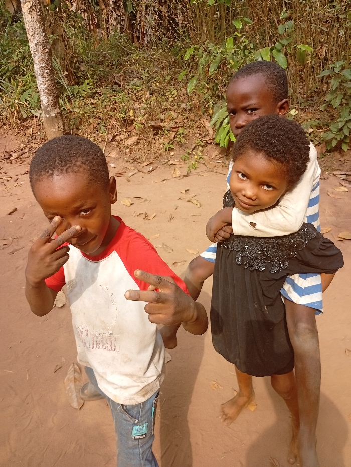 Three barefoot children are captured outdoors. A boy in a red and white shirt kneels, flashing a peace sign. He is wearing blue jeans with a patch. A girl with a black dress rides piggyback on another boy, who is wearing a blue and white striped shirt. The background consists of sparse vegetation, a tree trunk, and a sandy surface. All three children are looking directly at the camera.