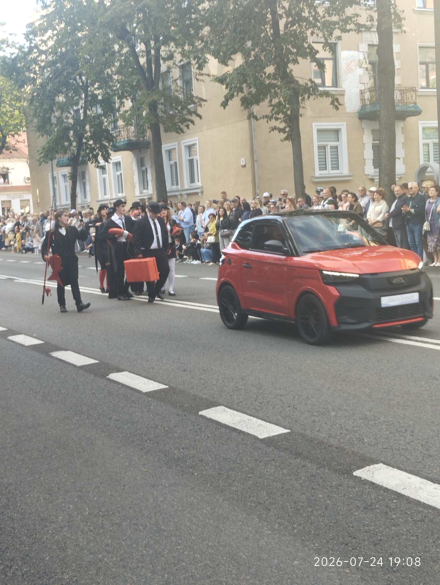 A group of performers in matching black suits and hats are walking in a procession on a street in Klaipėda, Lithuania. One performer is carrying a red prop that resembles a fishing rod, while another is carrying a bright orange suitcase. A small, red car with a black roof is driving alongside the group. The performers are on the road, with spectators lining the sidewalk in the background. Buildings with light-colored facades and numerous windows are visible behind the spectators, and trees with green foliage are interspersed among the buildings. The scene appears to be a parade or street event. The date and time "2026-07-24 19:08" are visible in the bottom right corner of the image.