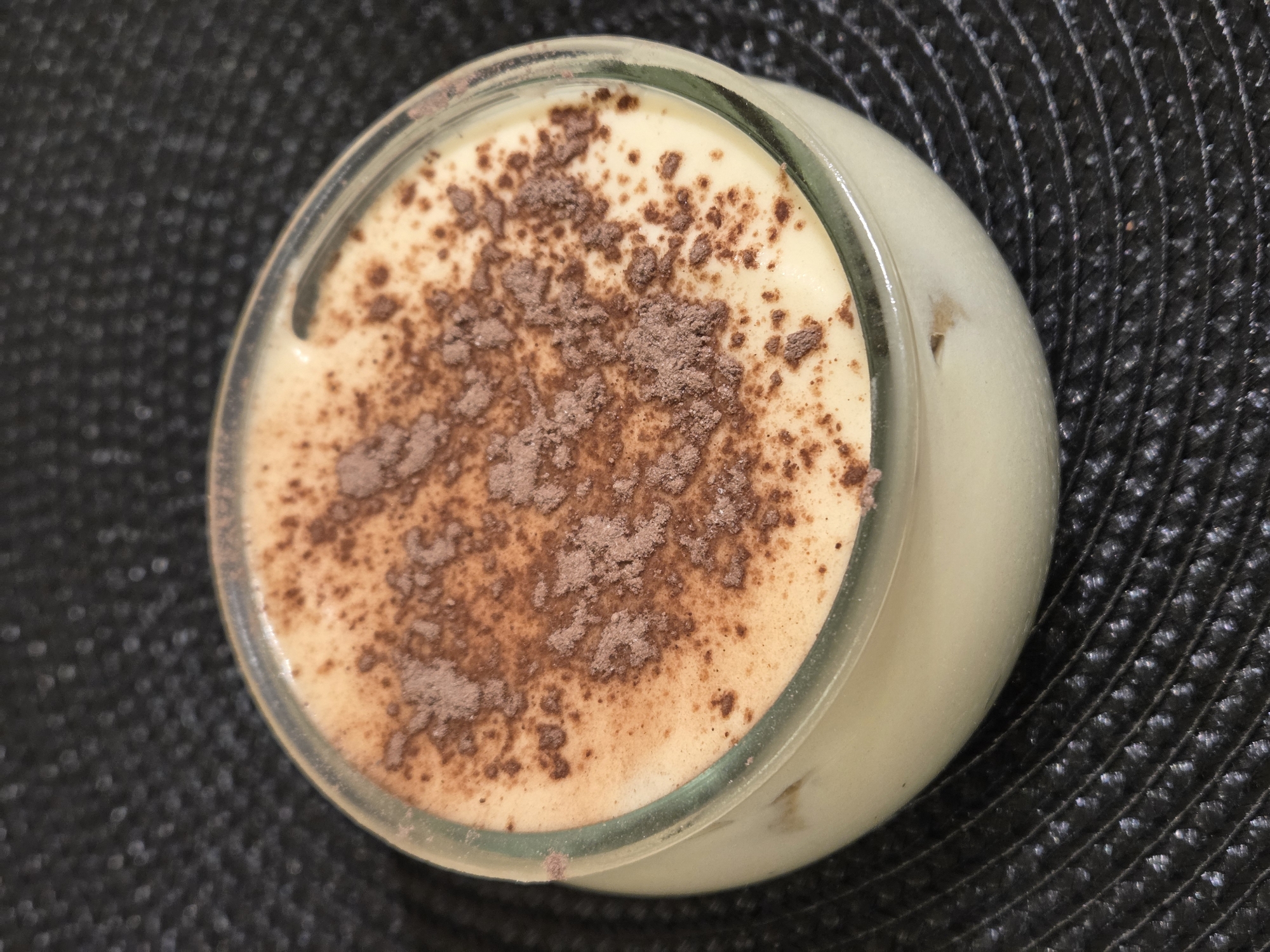 A close-up, high-angle shot shows a dessert in a glass jar on a textured, dark, likely black surface. The dessert has a light yellow or cream-colored base, topped with a scattering of dark brown powder, possibly cocoa. The jar is made of clear glass. The background shows the woven texture of a placemat or similar surface. There is no indication of this image being in Varna, Bulgaria.