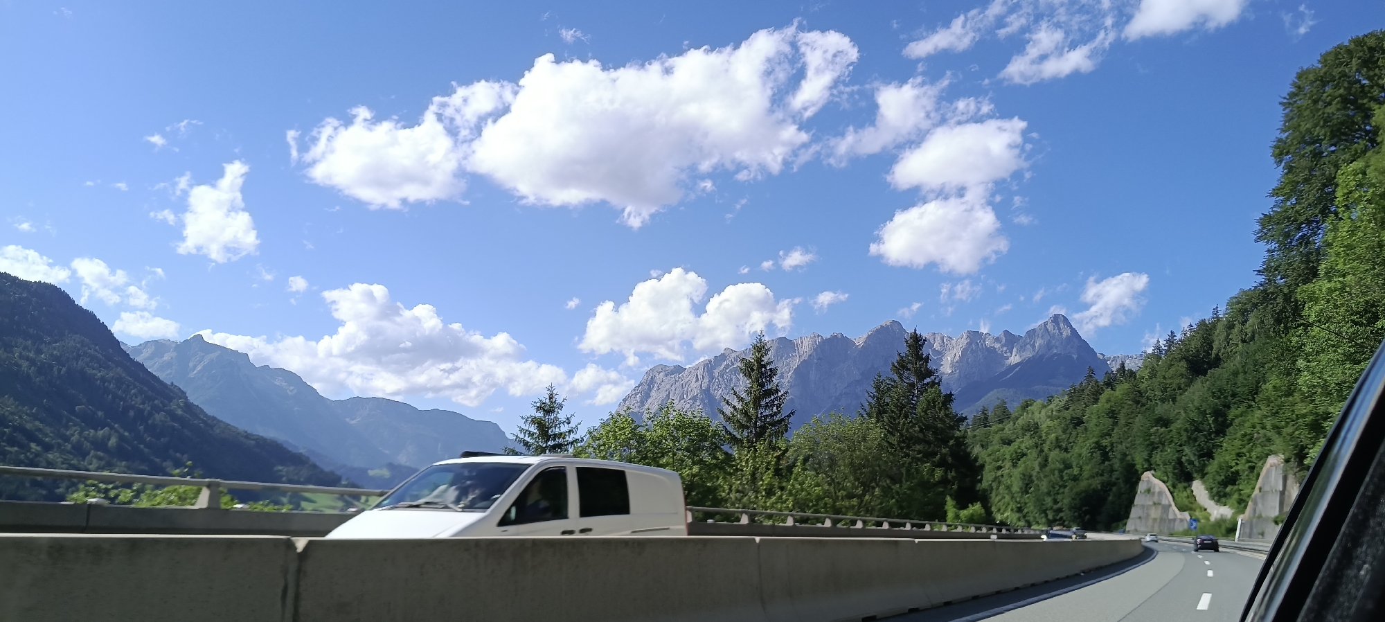 The image shows a view from a vehicle traveling on a highway with a concrete barrier in the foreground. To the left, a white van is visible. Beyond the highway, lush green mountains rise, with jagged, rocky peaks visible in the distance under a bright blue sky dotted with white clouds. The presence of mountains and the style of the highway suggest a mountainous region, possibly the Alps. While Bischofshofen, Austria is a city known for its mountainous surroundings and hosting ski jumping events, this specific scene cannot be definitively identified as being within the city limits of Bischofshofen without further contextual information.