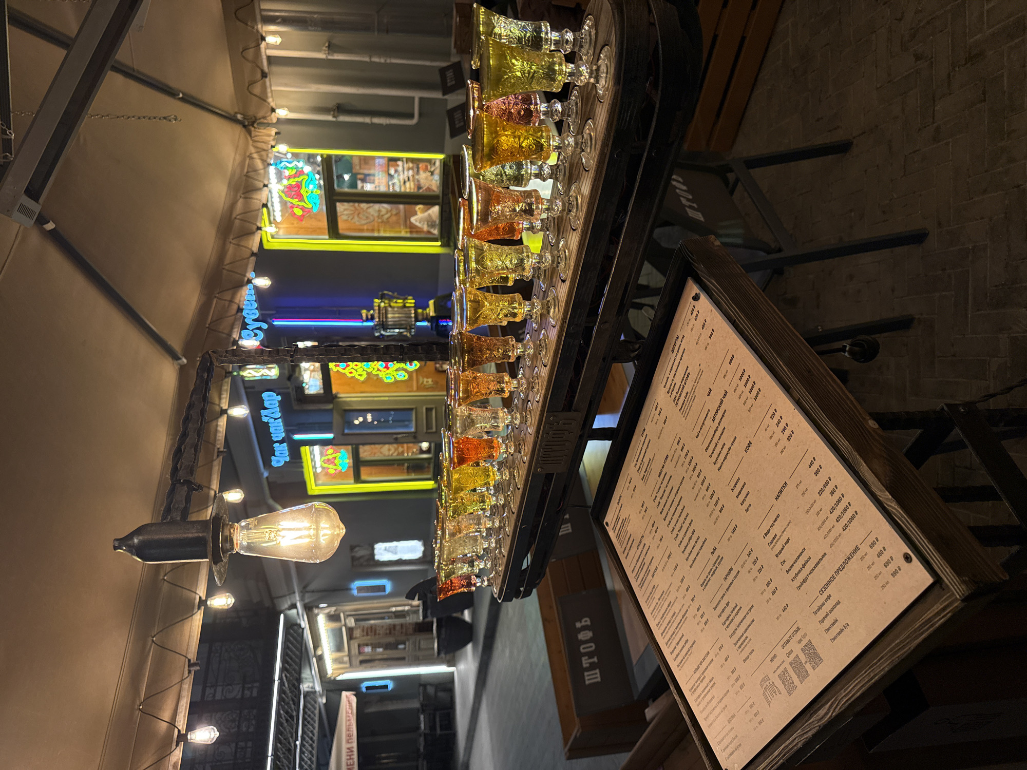 This photograph, likely taken at night, depicts a dimly lit outdoor dining area. The primary focal point is a bar top lined with numerous small glasses filled with colorful liquids, likely a selection of alcoholic shots or beverages. To the right of the glasses, a menu board, crafted from wood, displays menu items and pricing in Russian, including "ШТОФЪ" at the top. The table is positioned with a blurred and inverted view of an adjacent street, where businesses are identifiable by signage in Russian. Decorative lighting, in the form of string lights and a vintage-style lightbulb fixture, adds to the ambiance. Behind the bar, neon signs and decorative windows provide further illumination and contribute to the bustling atmosphere.
Given the presence of Russian signage and the menu content, the location is likely somewhere in Kazan, Russia.