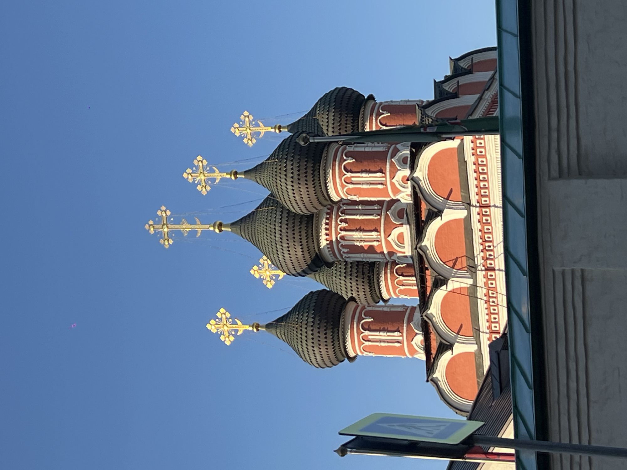 The image shows the Saint Basil's Cathedral in Moscow, Russia. The cathedral, with its distinctive red and white facade and colorful onion domes topped with golden crosses, is captured against a clear blue sky. The architectural details, including arched windows and decorative elements, are visible. In the foreground, a portion of a street sign with blue and white markings is visible on the left side. A green-tiled roof of a building is also in the lower part of the image, partially obscuring the base of the cathedral. There is a small, faint, purple object visible in the sky. The lighting suggests it is daytime, with sunlight illuminating the cathedral.