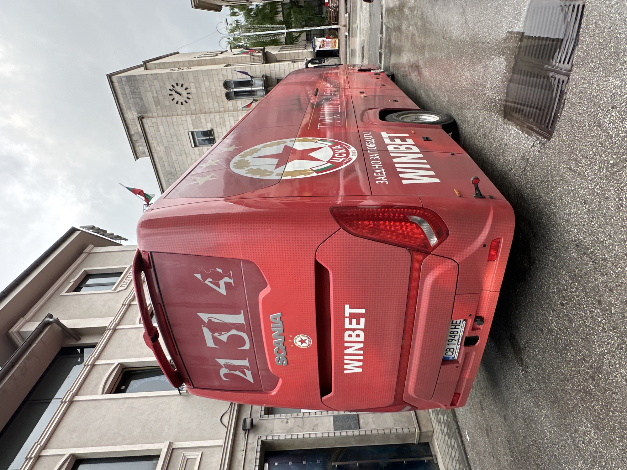 The image depicts a red SCANIA bus parked on a wet street in Veliko Tarnovo, Bulgaria. The bus is branded with "WINBET" and CSKA Sofia (a football club) logos, including the club's founding year "1948". The license plate number is "CB 1948 HE". The bus has the numbers "21" and "31" in white lettering. A building with a clock tower is visible in the background. The Bulgarian flag is visible on the bus. The street is paved with cobblestones.