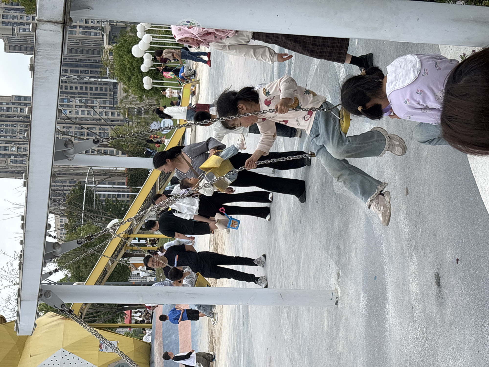 A wide shot of a playground in the city of Jiangjin, China. Several individuals, mostly children and adults, are present. Some are on swings, while others are standing nearby or in the background. The playground equipment includes swings suspended by chains from a structure with metal supports and a yellow component. In the background, multiple high-rise buildings are visible. The scene suggests an urban park environment with people engaging in recreational activities.