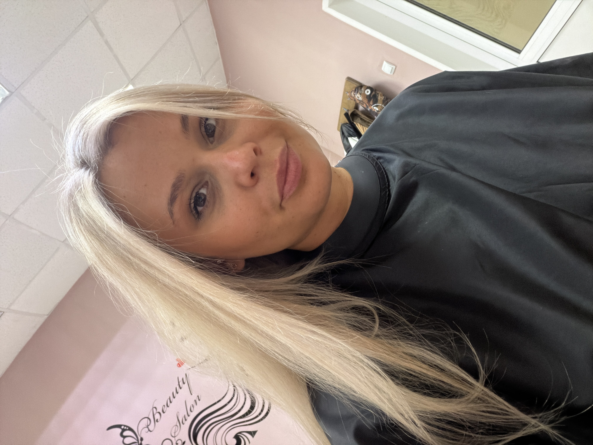 A medium shot shows a young Caucasian woman with long, blonde hair wearing a black cape in a beauty salon. The salon has light pink walls with a logo that reads "Beauty Salon" in cursive script. A window is visible in the background. The location is implied to be in Sarapul, Russia, based on the prompt's instruction.