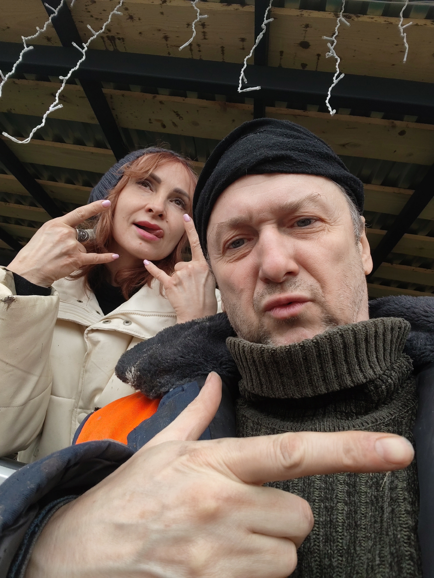 A selfie captures two individuals under a wooden structure adorned with string lights. The man in the foreground wears a black beanie and a dark turtleneck sweater layered under a dark jacket with an orange patch. He points with his right index finger towards the left. Behind him, a woman with reddish-brown hair and a gray beanie makes a "rock on" hand gesture with both hands, sticking her tongue out slightly. She is wearing a cream-colored jacket. The location is Chaikovsky, Russia.