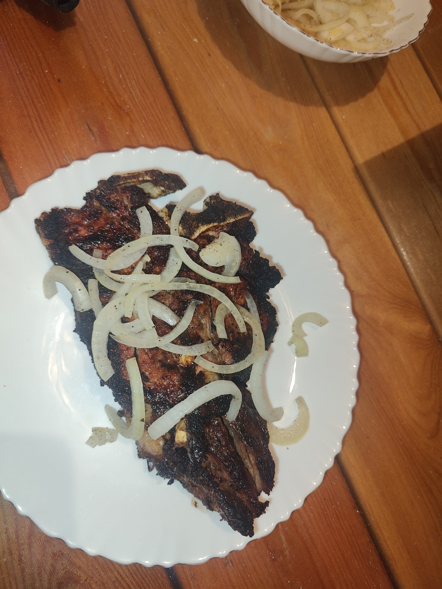 A close-up, overhead shot shows a plate of grilled meat with sliced onions on a wooden table. The meat is dark brown and appears to be ribs. The onions are white and thinly sliced, scattered over the meat. In the upper right corner, a white bowl with a gold rim contains more sliced onions. The wooden table has a visible grain and appears to be rustic. The lighting suggests an indoor setting. The image does not provide any information about the city of Izberbash, Russia.