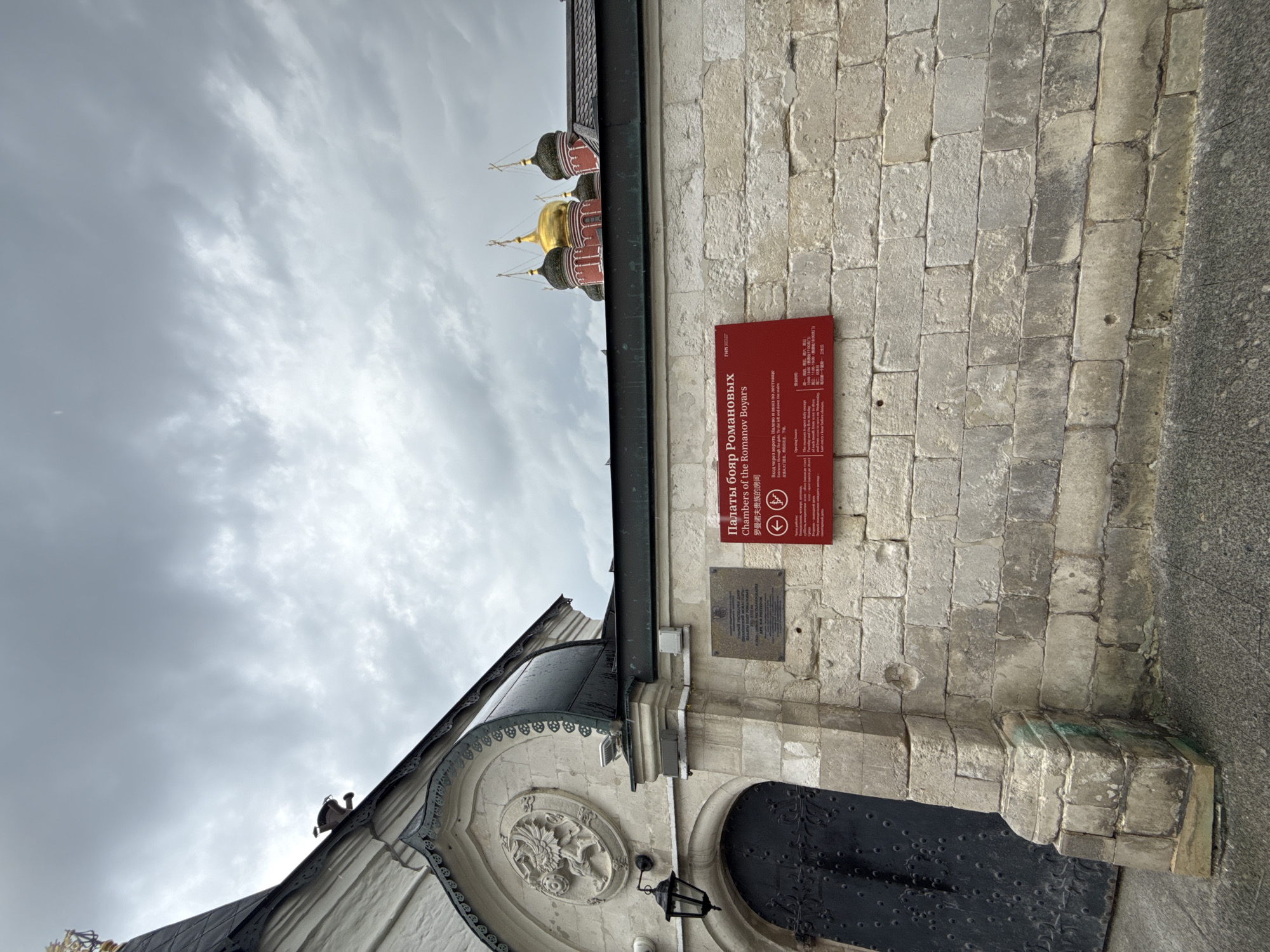 This image depicts a section of an old stone building, likely a historical landmark in Moscow, Russia. The building features ornate architectural details, including a carved circular medallion above a dark metal door. A black lantern hangs to the left of the door. Above the building, characteristic onion domes of a Russian Orthodox church are visible against a cloudy sky.
A prominent red sign is mounted on the stone wall. The sign displays text in Russian, English, and Chinese. The English text reads "Chambers of the Romanov Boyars," indicating the historical significance of the location as a residence of the Romanov family. The sign also includes operating hours, stating "11:00-19:00 (18:00)" and mentions "the first Monday of each month is a sanitary day."
The overall scene suggests a visit to a historical site, likely during the day given the light, though the cloudy sky implies overcast weather. The stone construction of the building suggests a long history, and the presence of multiple languages on the sign indicates it is a tourist attraction. No people are visible in the frame.