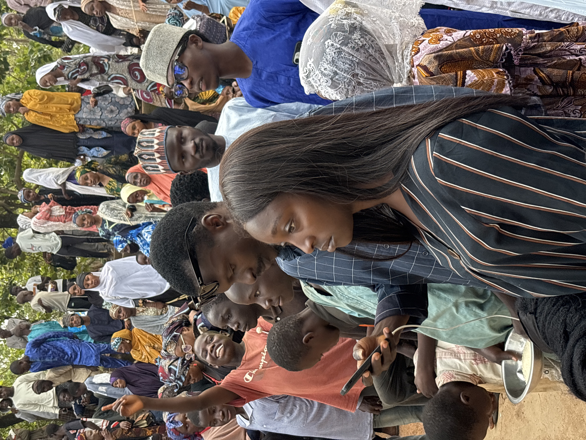 The image depicts a gathering of people in an outdoor, earthy environment, likely in Jos, Nigeria. The scene is filled with individuals of varying ages and genders, many of whom are wearing traditional African attire, including vibrant fabrics and head coverings.
In the foreground, a group of people are closely packed together. A young man in a blue checkered shirt is looking towards a smartphone in his hand, which is connected to a white charging cable. Next to him, a woman with long dark hair is visible from the side, also looking downwards. To their left, a boy with a red t-shirt is looking up and smiling, with his arm raised. He appears to be holding a small object in his hand. A man wearing glasses and a traditional hat is in the center, looking towards the right. Another man, also in traditional attire and a hat, is visible behind him.
In the background, a larger crowd is visible, with many individuals standing or sitting. There is a variety of clothing styles, indicating a diverse group. Some are wearing brightly colored outfits, while others are in more subdued colors. The environment suggests a rustic setting, possibly a village or a park, with trees and natural ground visible. The overall impression is of a community event or gathering.