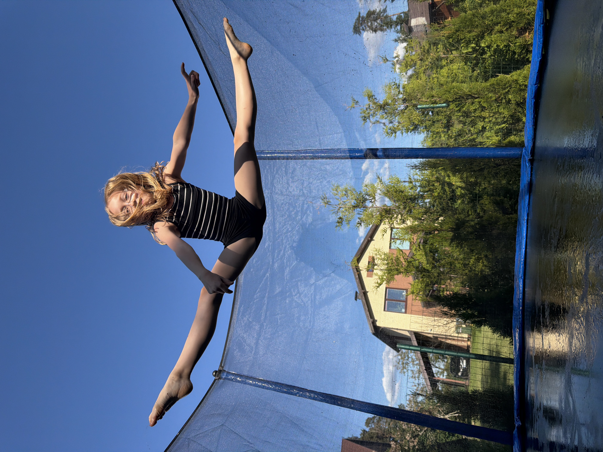 A young female individual with blonde hair is captured mid-air above a trampoline, performing a straddle split with outstretched arms. She is wearing a black and white striped sleeveless top and black bottoms, with bare feet. The trampoline features a blue safety net enclosure supported by blue poles, with a wet black mat and blue padded spring cover visible in the foreground. In the background, under a clear blue sky with scattered white clouds, a two-story residential house with light yellow/tan walls and dark windows is partially visible. Green coniferous trees and bushes surround the house and are positioned between the trampoline and the residence. A fence and a dark-colored vehicle under a carport are discernible through the foliage. The scene depicts an outdoor, sunny environment in Dreiliņi, Latvia.