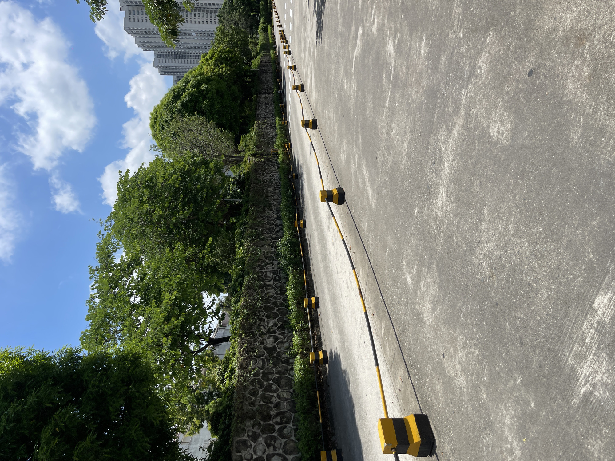 A sunny day with a blue sky and scattered clouds, the image shows a paved road lined with yellow and black traffic delineators. A stone wall with lush green foliage growing on top borders the road on the left. Beyond the wall, trees are visible. In the background, modern high-rise buildings stand tall. The road appears to be a pathway or lane, possibly within a residential or commercial area in Panyu District, China. There are no people or vehicles present in the scene.