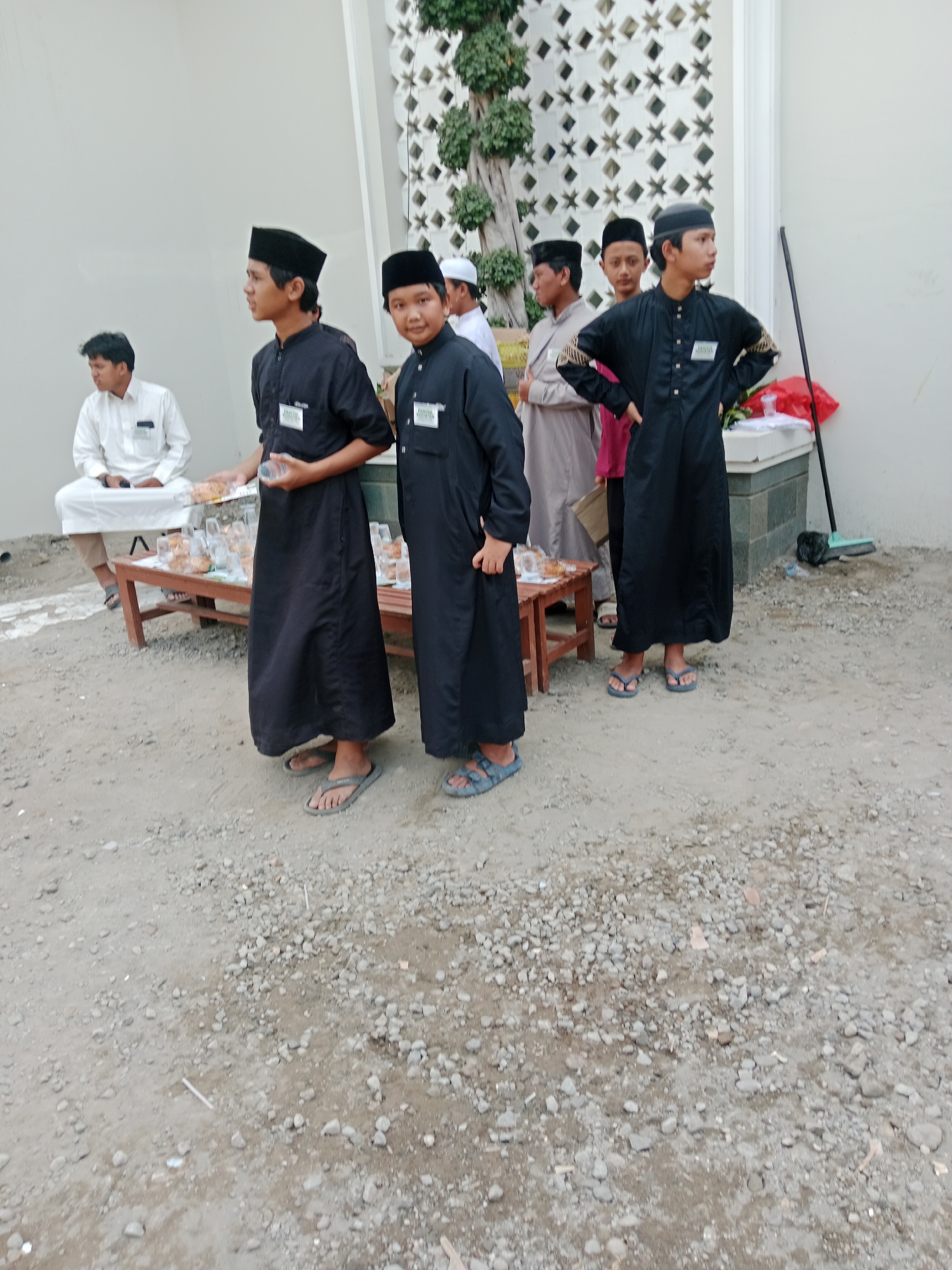 In the city of Kediri, Indonesia, a group of young men are gathered outdoors. The scene appears to be near a building with a decorative white wall and a tree. Several of the men are wearing traditional Islamic attire, including black caps and black robes. One man in a white robe is seated on a bench. A wooden bench with trays of food and plastic cups is set up in the foreground, suggesting an outdoor event or gathering. The ground is covered with gravel.