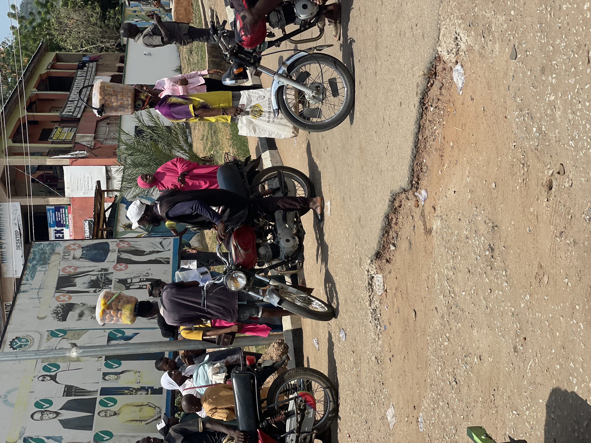 Here is a detailed description of the image:
The image appears to be taken on a sunny day in an urban area with a number of people present. The location features a building visible in the background.
Several people are standing near motorcycles, which are parked alongside a paved road or street. Many are either on or around the motorcycles. A person carries a container on their head.
There's an advertising poster behind the group, with images and text including the phrases "Superior Quality," "vital," and details related to training and monetary transactions like "deposit," "withdraw money," "send money," and "buy airtime." Also, there is the phone number 2,07065589766. The poster also contains portraits of people with check marks and crosses on their images.
The ground adjacent to the paved area appears to be a dirt surface with debris. The building in the background has signage that reads "FEDERAL IMPRESSION ST."