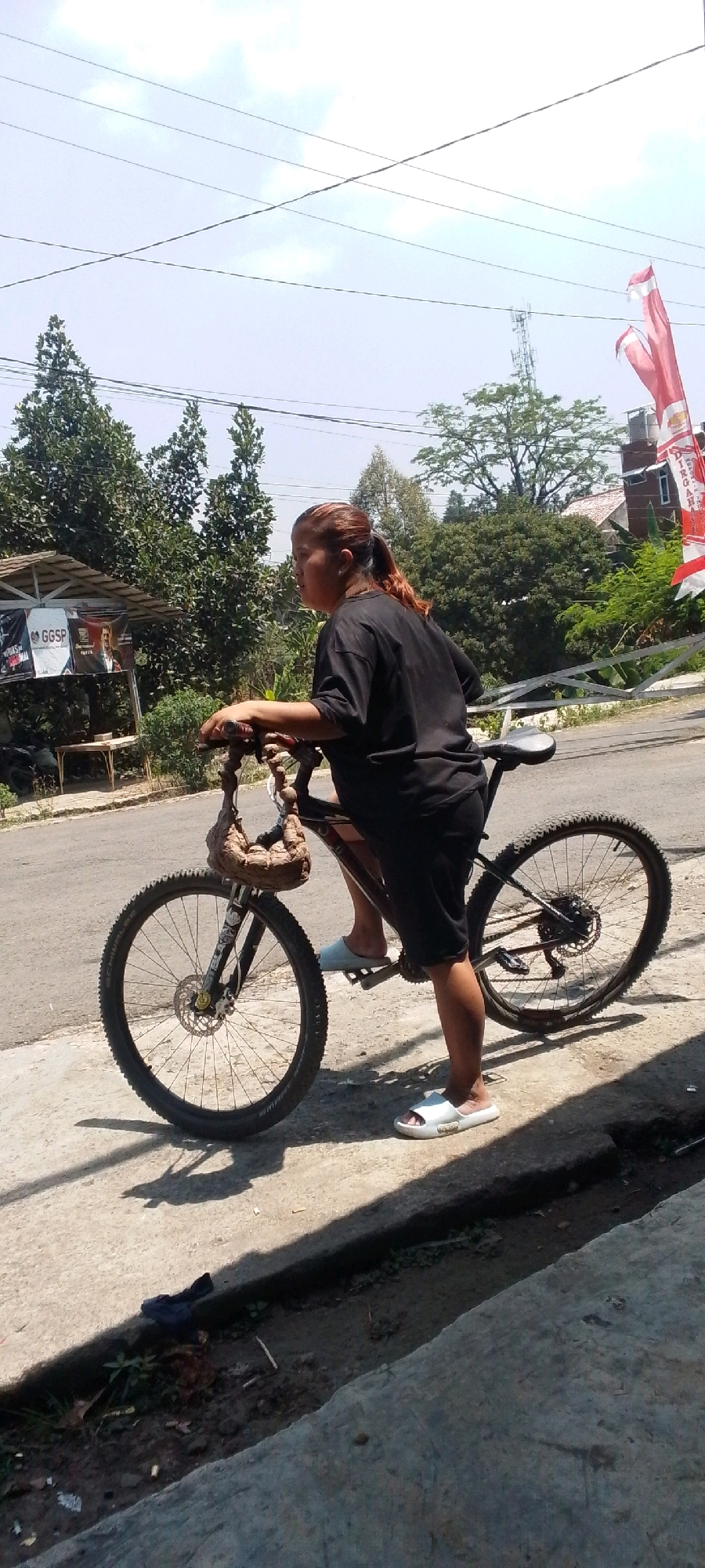 In Cilaku, Indonesia, a woman in a black t-shirt and shorts stands beside a bicycle. The bicycle has a basket attached to the handlebars, and the woman is holding the handlebars. She is wearing white sandals. The scene is outdoors, with a road and sidewalk visible. Trees and some buildings are in the background. The sky is clear and blue, with some power lines overhead. A red and white flag is also visible in the background. The woman appears to be looking off to the side.