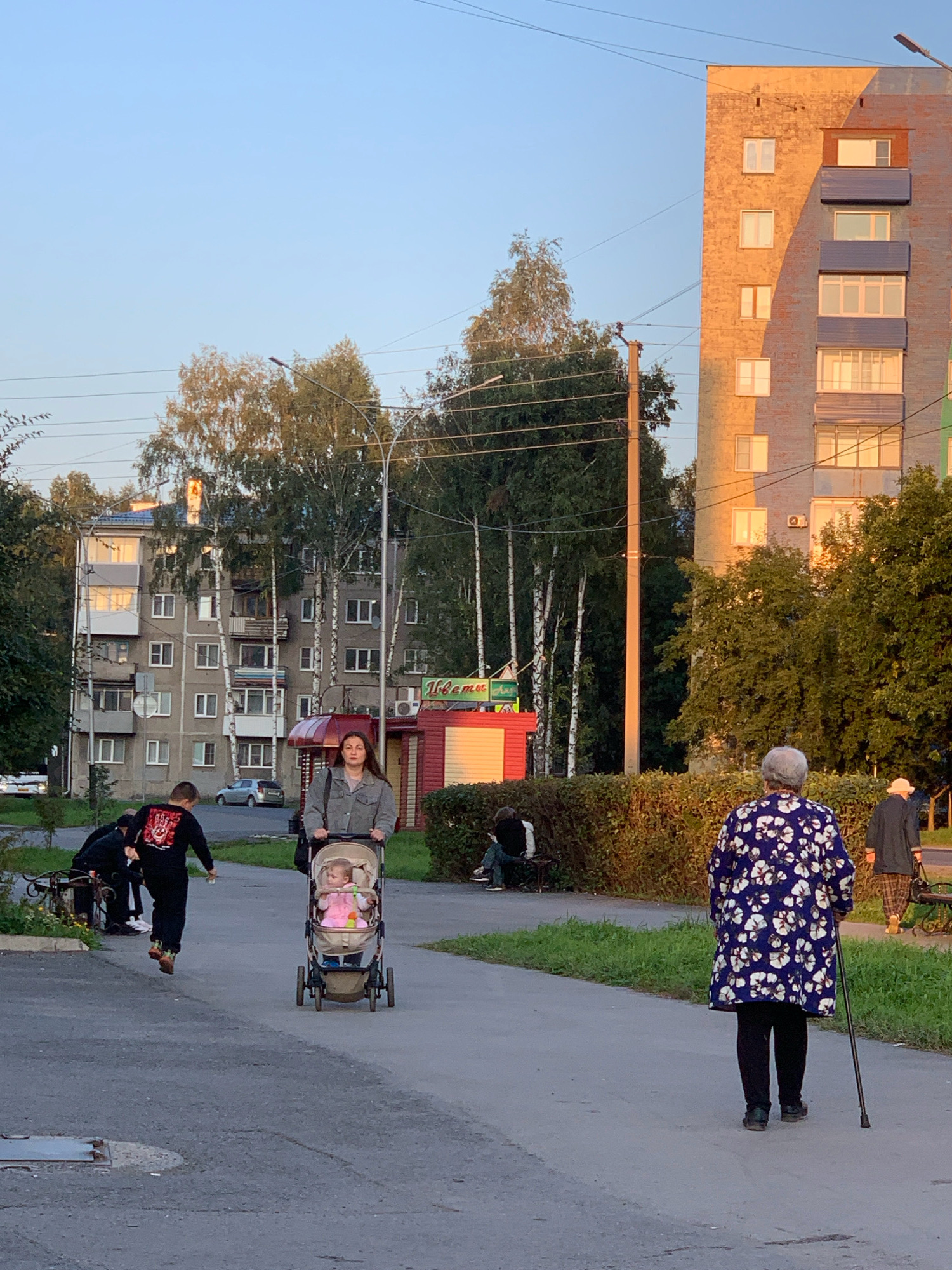 The image depicts a street scene in Prokopyevsk, Russia, on a sunny afternoon. In the foreground, a woman is pushing a stroller with a baby inside. To her left, a young boy is running. In the background, an older woman with gray hair is walking with a cane. Further in the background, there are residential buildings, trees, and a small kiosk. The sky is clear and blue, and the sun is casting long shadows, indicating it is late afternoon. The overall impression is a typical urban environment with people engaged in daily activities.