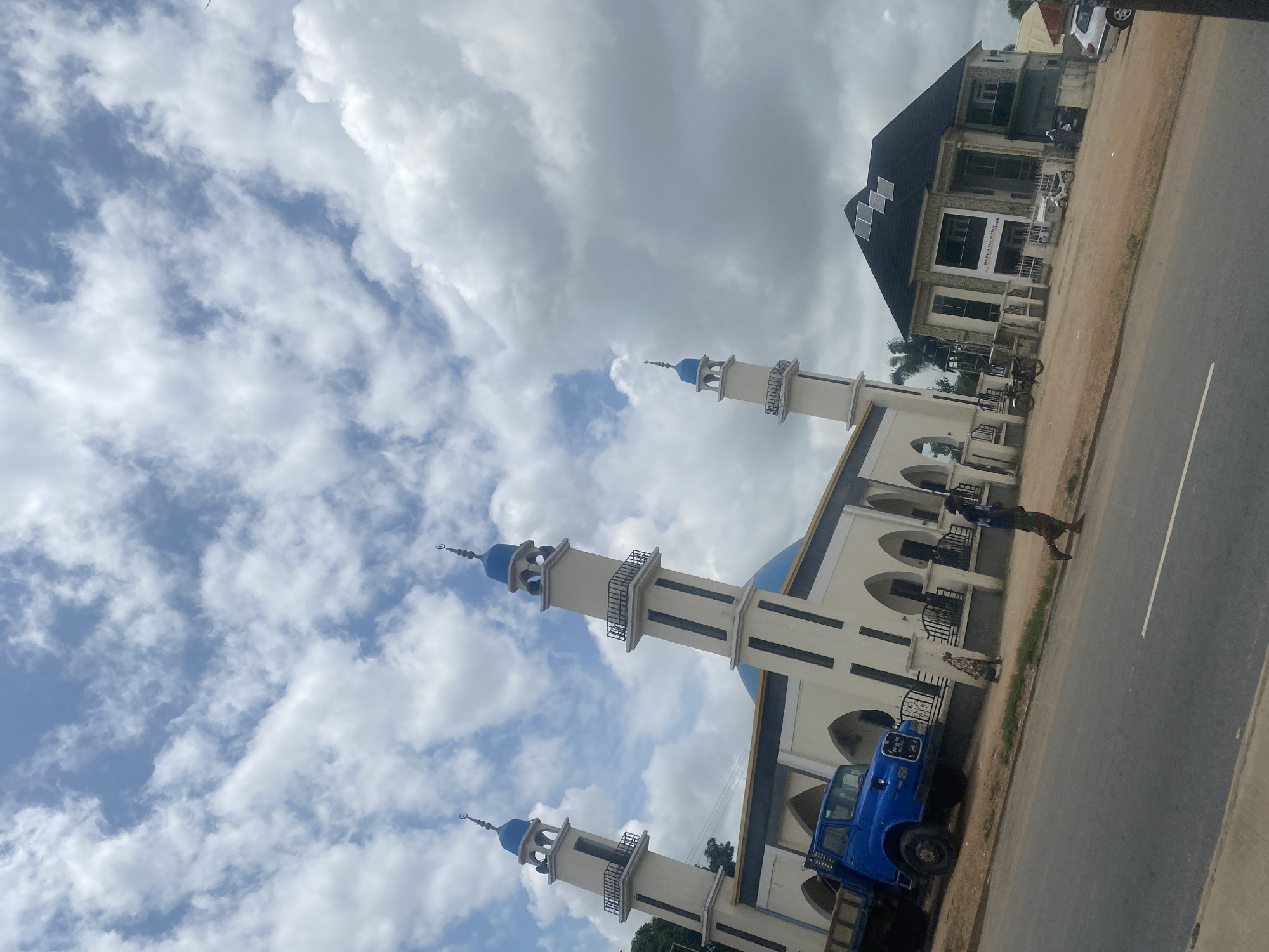 This image, taken in Gudi, Nigeria, shows a mosque with two prominent minarets, each topped with a crescent moon. The mosque features a large blue dome and white walls with arched windows and doorways. In front of the mosque, a blue truck is parked. To the right of the mosque is a modern building with a dark roof and solar panels, and a sign that reads "BARKA DA ZUWA". A person is walking on the road in front of the mosque, and another person is walking in the background. The sky is filled with dramatic clouds.