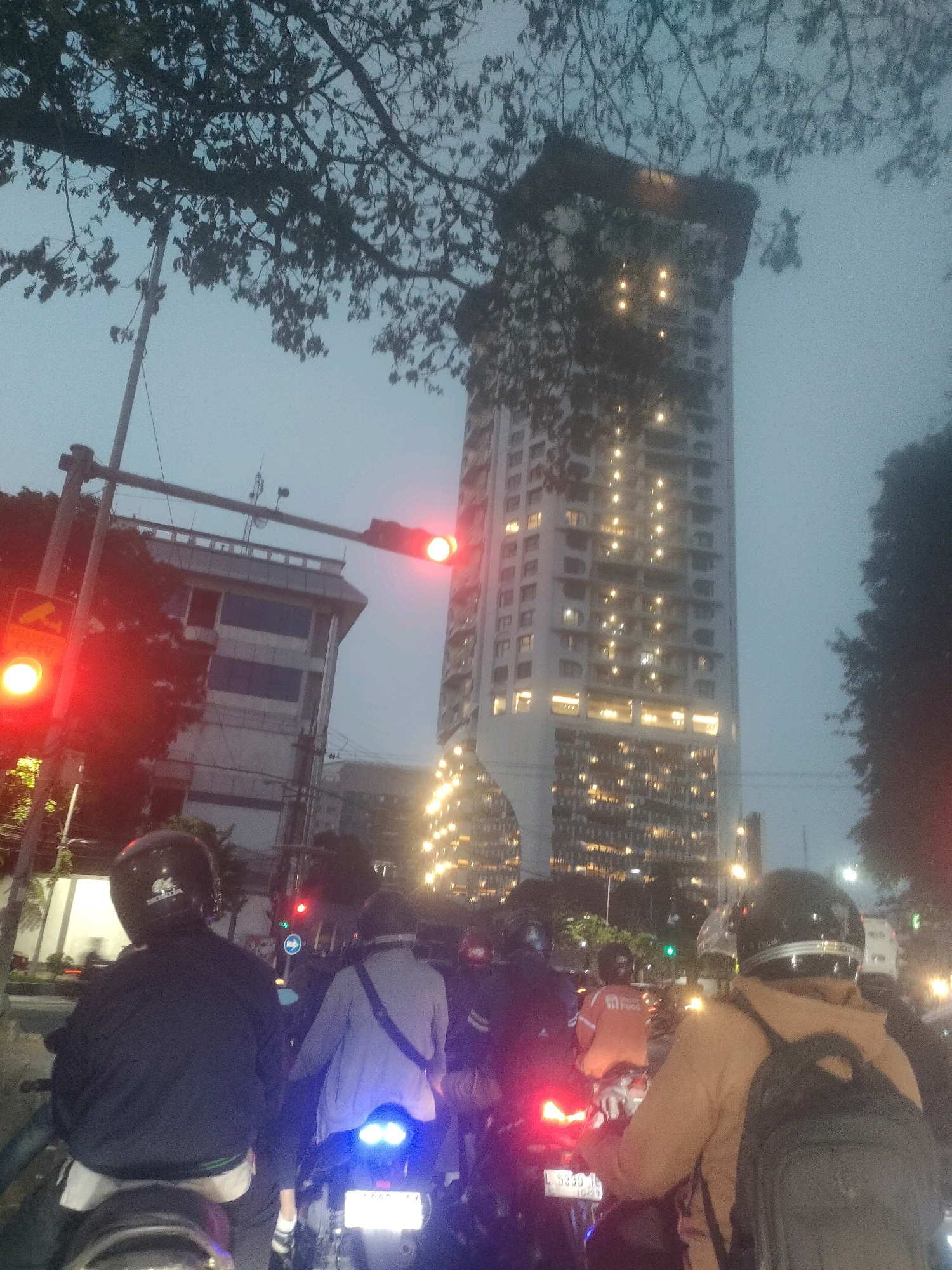 The image depicts a street scene in Surabaya, Indonesia, during dusk. A tall, modern apartment building with many illuminated windows dominates the background, suggesting an urban setting. In the foreground, several people on motorcycles are stopped at a traffic light, which is currently red. The cyclists are wearing helmets and casual clothing. One motorcyclist in the immediate foreground wears a black jacket and a black helmet, and is seen from behind. Another motorcyclist in front of them wears a light blue jacket and is also seen from behind. A third motorcyclist to the right, also seen from behind, wears a tan jacket and a black backpack. The traffic light is positioned over the road, and a traffic sign is visible to the left of the light. Trees with sparse foliage partially obscure the view of the buildings and the sky, which is a muted gray. The overall atmosphere is that of a typical evening traffic situation in a busy city.