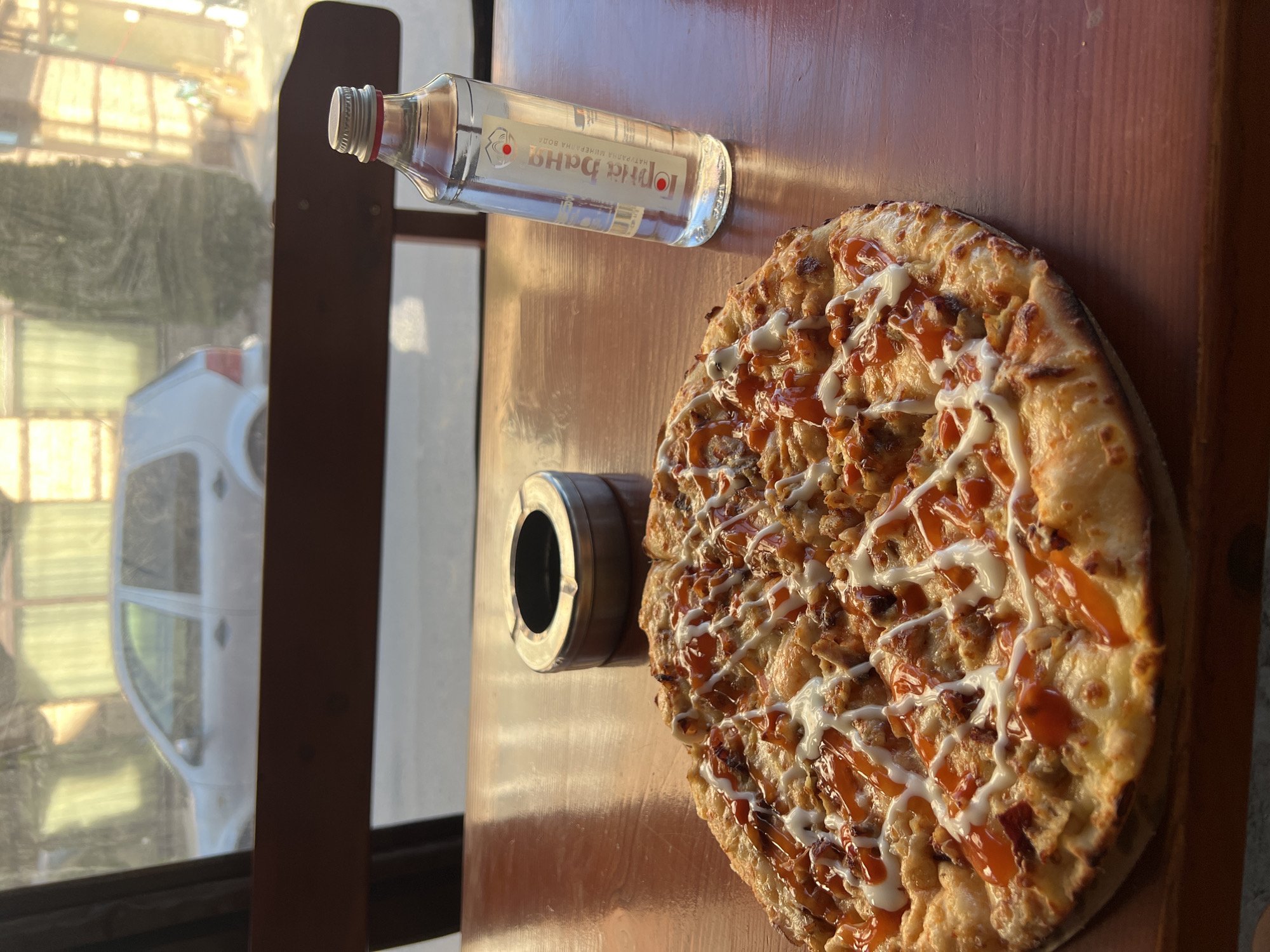 On a wooden surface, likely a table, are arranged a pizza, a small glass bottle, and a metal ashtray. The pizza is round, topped with what appears to be meat or vegetables, and drizzled with white and orange sauces. The glass bottle is small, possibly containing a liquid, and features a label in a non-English script. The metal ashtray is cylindrical. Outside a window, a glimpse of a white car and greenery suggests an exterior setting. The scene is illuminated by daylight. The information about Breznitsa, Bulgaria is not evident in the image.