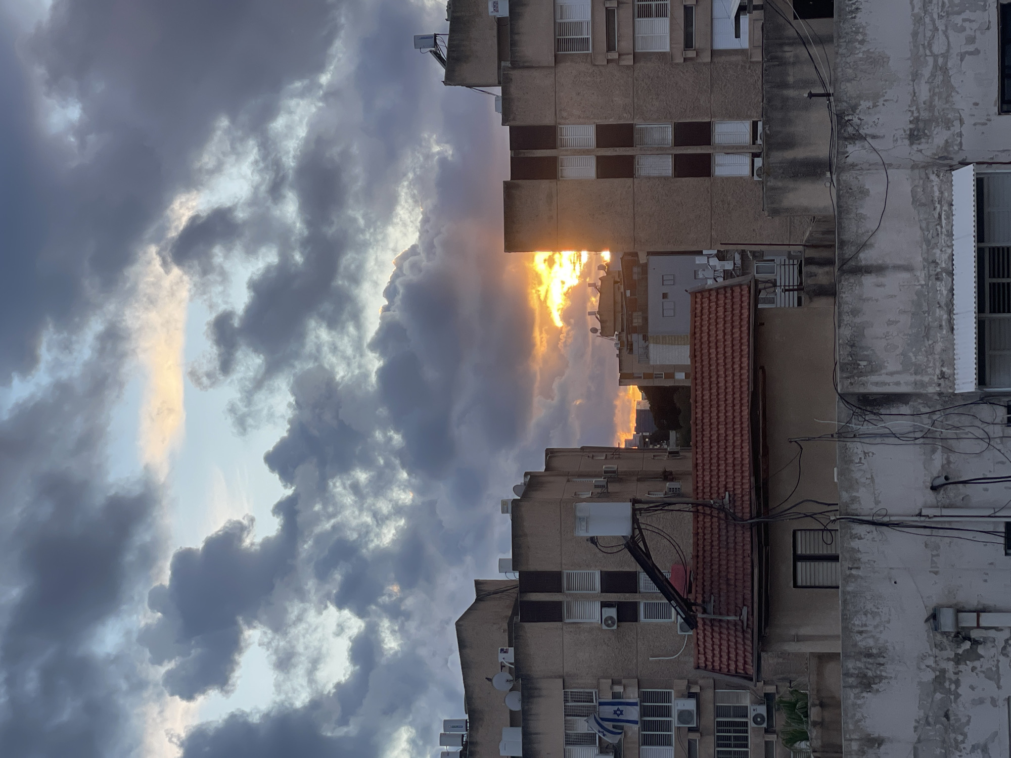 This image depicts an urban residential area in Petah Tikva, Israel, at either sunrise or sunset. The sky is largely overcast with dense, dark grey cumuliform clouds. A prominent orange-yellow sunburst is visible on the horizon between two buildings, casting a bright glow and illuminating the undersides of some clouds.
In the foreground, a weathered, light-colored concrete or stucco wall runs horizontally, showing discoloration and patches of peeling surface. Multiple black electrical cables and wires are strung haphazardly across and down this wall. Below the wall on the right, a white awning is partially visible.
Behind the foreground wall, a smaller, light-brown building with a distinctive red-tiled gabled roof is present. Additional black electrical wiring is draped over this roof and extends to other structures. A dark, rectangular window is set into its facade.
To the left, a multi-story residential building, constructed from light-brown stucco, occupies a significant portion of the frame. It features numerous rectangular windows, many equipped with white external louvered shutters. Multiple white external air conditioning units are mounted on its facade. A blue and white flag of Israel hangs from a lower balcony. Its flat roof supports several white cylindrical objects, likely solar water heaters, and multiple satellite dishes. A small dark bird is perched on the roof's edge.
To the right, another similar multi-story stucco building with white louvered windows and visible air conditioning units stands. Its roof also displays utilities, including what appears to be a solar water heater.
Distant residential buildings with less defined features are visible on the horizon, partially obscured by the atmospheric light from the sun. No human figures or specific actions are discernible. The scene presents a typical urban landscape with dense housing and visible utility infrastructure under a dramatic, cloudy sky.