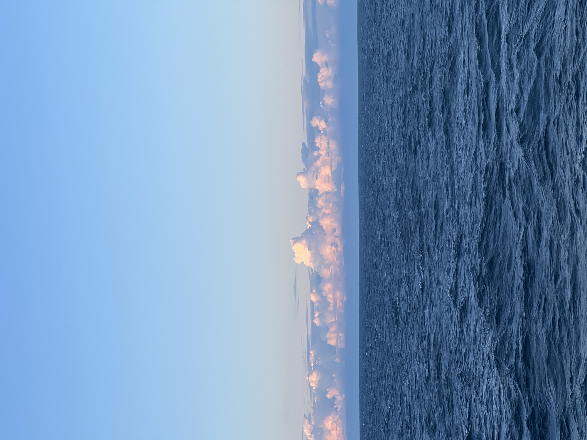 An expansive, clear blue sky dominates the upper portion of the image. Below the sky, a band of fluffy white clouds, tinged with pink and orange from what appears to be sunrise or sunset, stretches across the horizon. Beneath the clouds, a vast expanse of deep blue ocean fills the lower half of the image, with small, choppy waves creating texture on its surface. The scene depicts a serene seascape under a colorful sky.