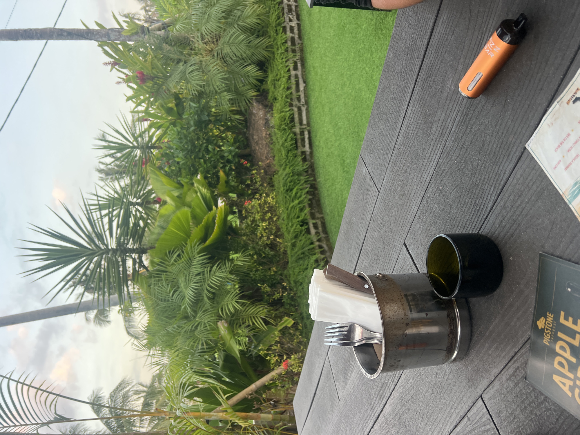 This is a medium shot, eye-level outdoor photograph of a table setting with a lush tropical backdrop. The table surface is made of dark gray wooden planks. On the table, a silver metal bucket holds two forks and a stack of white napkins, with a small dark green glass cup to its right. In the foreground, a menu with the words "PIGSTONE TAPHOUSE APPLE CID" is visible. An orange electronic cigarette device rests on the table further to the right. Behind the table is a vibrant green lawn, bordered by a low stone wall. Beyond the lawn, a dense arrangement of tropical plants and palm trees creates a lush, green backdrop under a cloudy sky. There are no people clearly visible in the frame. The scene suggests a casual dining or drinking establishment in a tropical location. The image does not contain any elements that would allow for identification of the city of Kediri, Indonesia.
