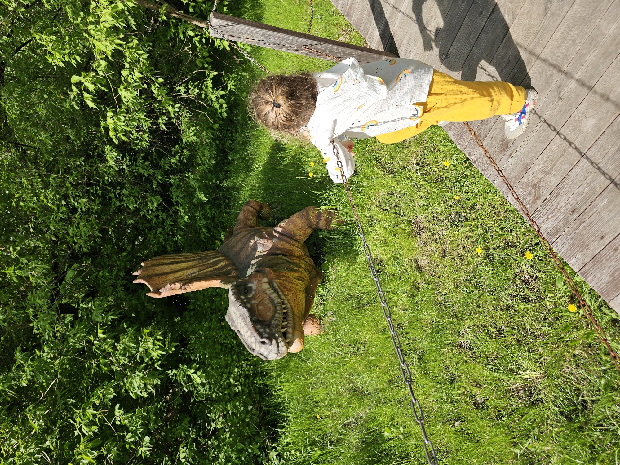 A child stands on a wooden walkway next to a chain railing overlooking a lawn with a dinosaur statue. The child has brown hair pulled back in a small ponytail, wears a white shirt with rainbows, and yellow pants. The dinosaur is brown and green with a gray head and large teeth. The lawn is green with some dandelions. Green bushes surround the lawn. The location is Khimki, Russia.