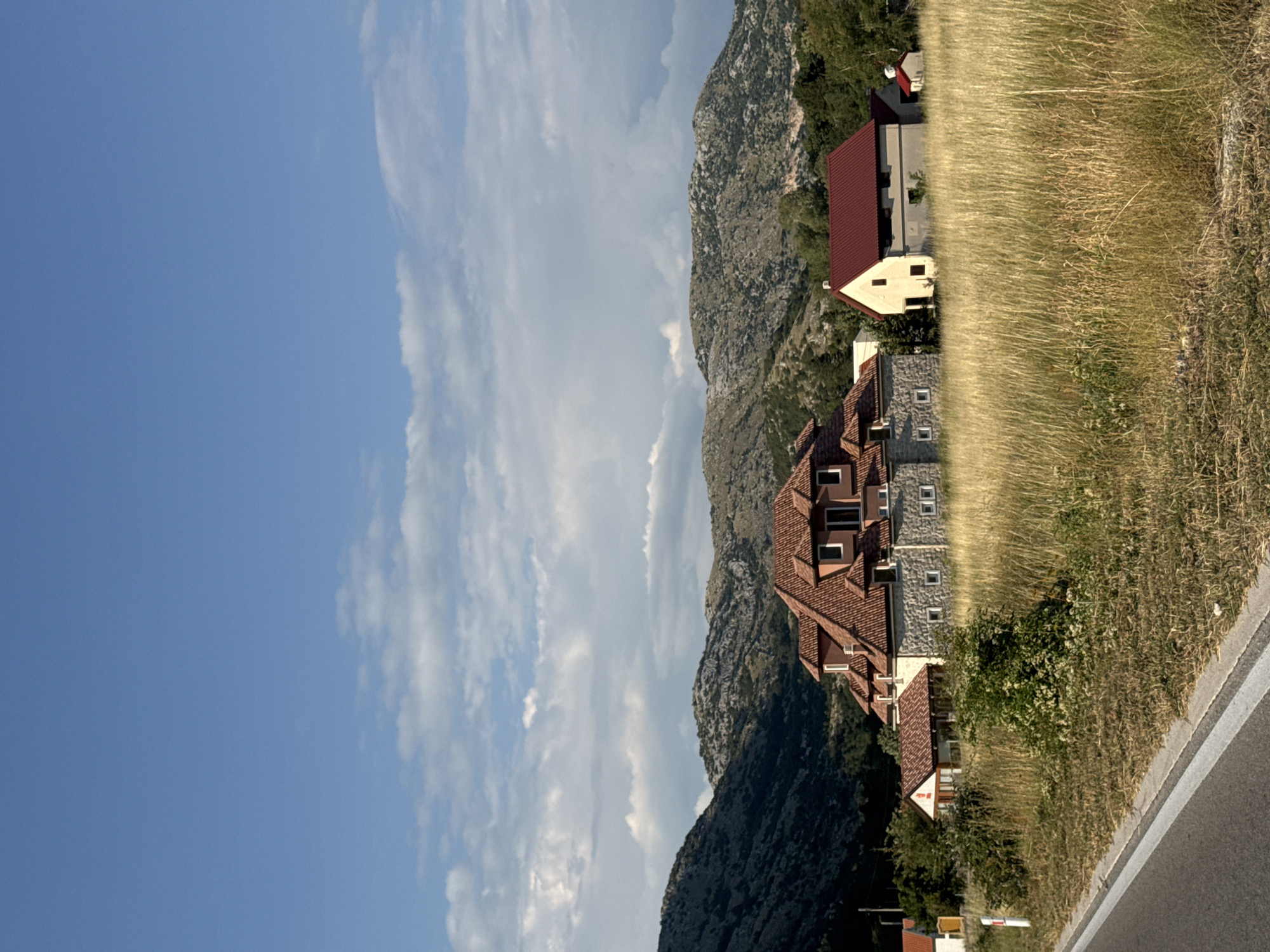 The image captures a rural scene in Kopito, Montenegro, under a bright blue sky with scattered white clouds. In the foreground, a dry, tall grass field stretches out, bordered on the left by a paved road with a white line marking its edge. A small white marker with a red top stands near the roadside.
Dominating the middle ground are several buildings. The largest is a substantial house with a multi-tiered, reddish-brown tiled roof and stone-clad lower walls. It has several windows visible. To its right, a smaller, simpler building with a red roof and pale yellow walls is situated. Further to the right and behind this, a tiny red-roofed structure is visible. Green trees and shrubs dot the landscape around the buildings.
In the background, rugged, rocky hills rise up, covered in sparse vegetation. The terrain suggests a mountainous or hilly region. The lighting indicates it is likely daytime, possibly late afternoon given the long shadows. No people are visible in the image.