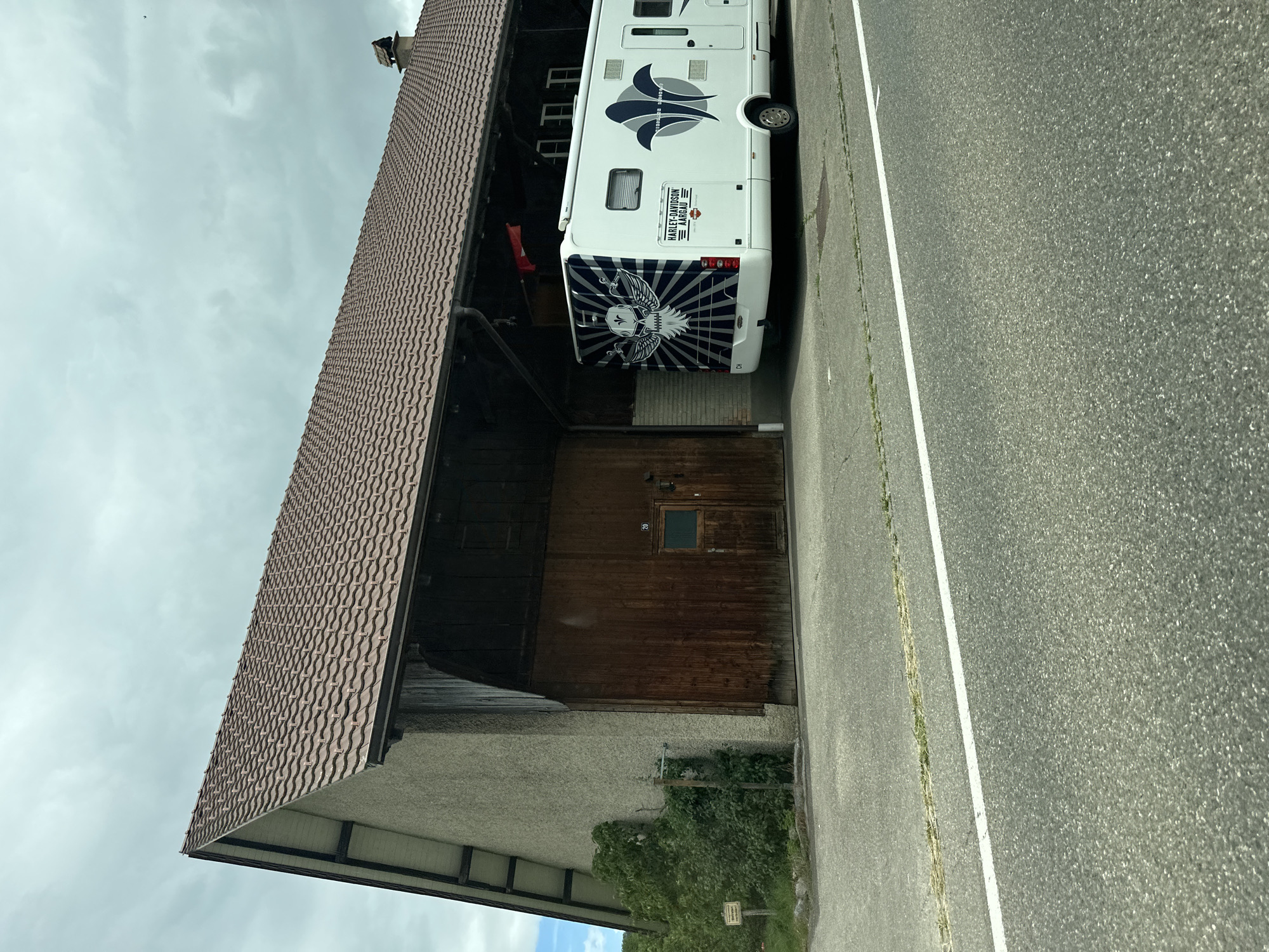 The image captures a white recreational vehicle (RV) parked alongside an asphalt road, next to a large building, under an overcast sky. The scene is set in Zurzach, Switzerland.
The RV is prominent, displaying extensive custom graphics. On its rear, a large blue and white design features an eagle-like bird with spread wings holding a skull or crest, against a sunburst background. Above this, a white rectangular plaque reads "HARLEY-DAVIDSON AARGAU," and a "CH" country sticker (indicating Switzerland) is visible below the eagle graphic. The side of the RV has an abstract blue and grey design.
To the left of the RV stands a substantial building, possibly a barn or agricultural structure, with a prominent brown, tiled gabled roof. The upper section of the building is constructed from dark wood, with small windows. Its lower part features large wooden garage doors, one of which has a small rectangular window. The base of the building is composed of a lighter-colored, textured wall, with some green bushes and foliage growing nearby.
An asphalt road occupies the right side of the frame, marked by a clear white solid line along its edge. Patches of grass and weeds are visible growing along the roadside. The sky above is uniformly grey and cloudy, indicating an overcast daytime. The RV appears to be parked or stored, with no human activity immediately apparent.