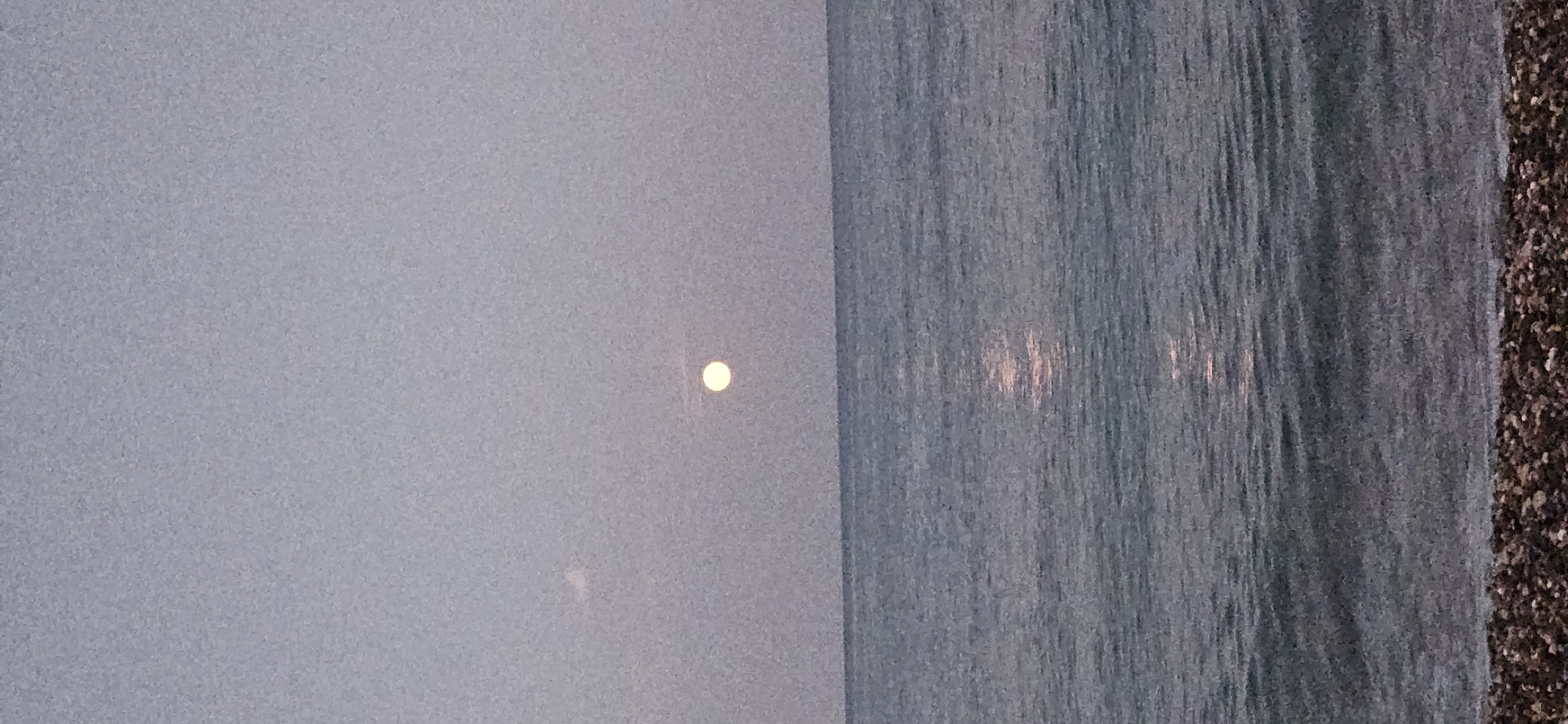 The image shows a serene twilight scene at a beach in Kemer, Turkey. The sky is a muted gray, with a soft, pale full moon visible in the upper center of the frame. A faint reflection of the moon shimmers on the calm, blue sea. The water gently laps at a pebbled shoreline in the foreground, suggesting a quiet, uncrowded beach. The overall atmosphere is peaceful and tranquil, characteristic of a sunset or early moonrise along the Mediterranean coast.