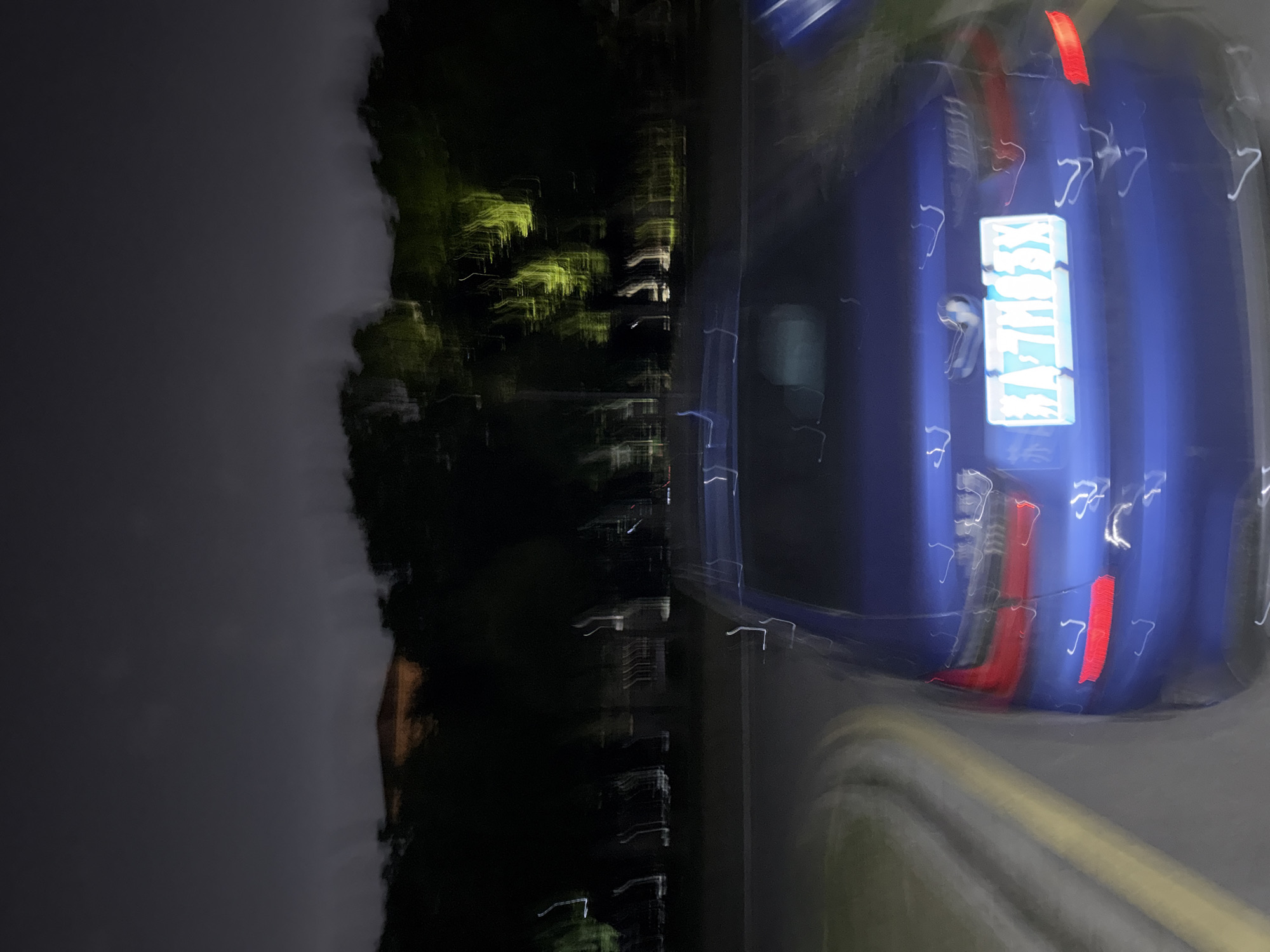 The image depicts a blurred scene at night, likely taken with a moving camera. A blue vehicle is partially visible on the right side of the frame, its taillights illuminated. To the left, there's a dark area suggesting trees or vegetation. There's also a sign on the vehicle, but the text is illegible due to the blur. The location is believed to be in the city of Jiangning District, China. Overall, it's a night scene with a focus on the vehicle and blurred surroundings.