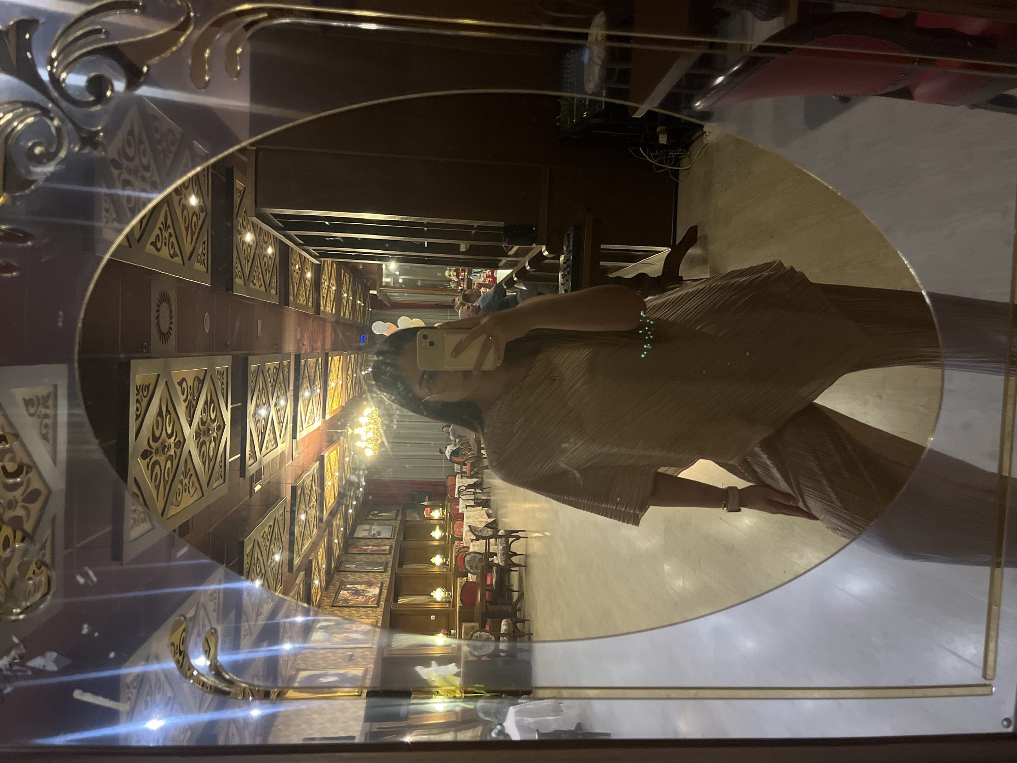 The image shows a reflection in a large, ornate mirror. The mirror is framed with gold and dark wood details. A person is visible in the reflection, holding a phone and appearing to take a selfie. They are wearing a brown or tan pleated outfit and jewelry.
The reflection shows a long, grand room, likely a hotel or restaurant interior. The room features ornate walls with decorative panels or artwork, and chandeliers hanging from the ceiling. There are tables and chairs set up in the room, suggesting a dining area or event space. The overall style suggests a luxurious or historical setting. The specific location and city cannot be determined from the image alone.