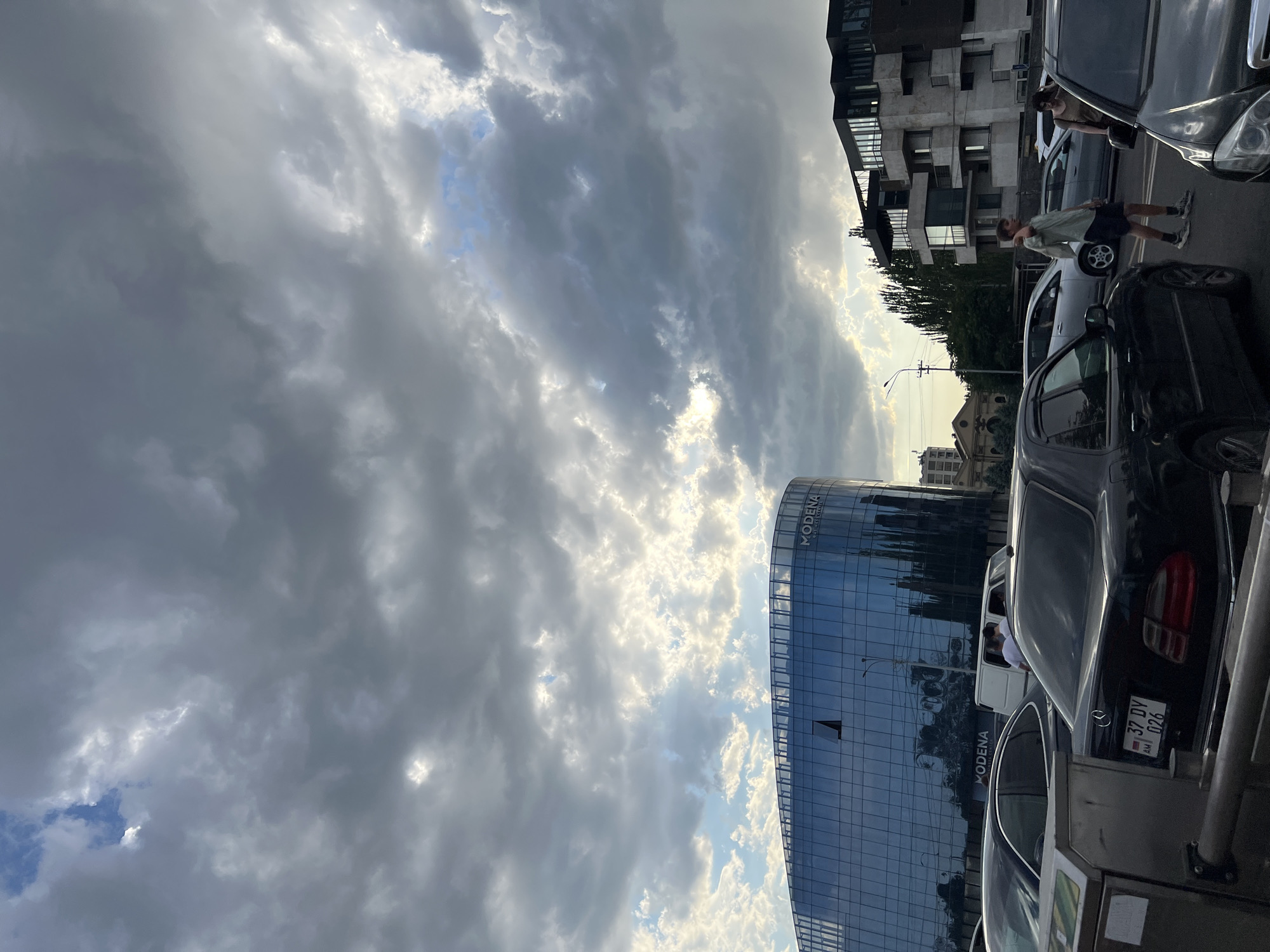 The image captures a street scene in Yerevan, Armenia, with a prominent curved glass-fronted building labeled "MODENA BUSINESS CENTER" in the background. The sky is filled with dramatic, partly cloudy formations, with patches of blue sky and sunlight breaking through. Several cars are visible, including a black Mercedes-Benz sedan in the foreground with a license plate reading "37 DV 026". A white van and other vehicles are also present, suggesting moderate traffic. Two individuals are walking on the sidewalk or crossing the street near the Modena building. The architecture of the buildings appears modern and somewhat varied, with the Modena building being the most striking. The overall atmosphere suggests a late afternoon or early evening setting, given the lighting and cloud cover.