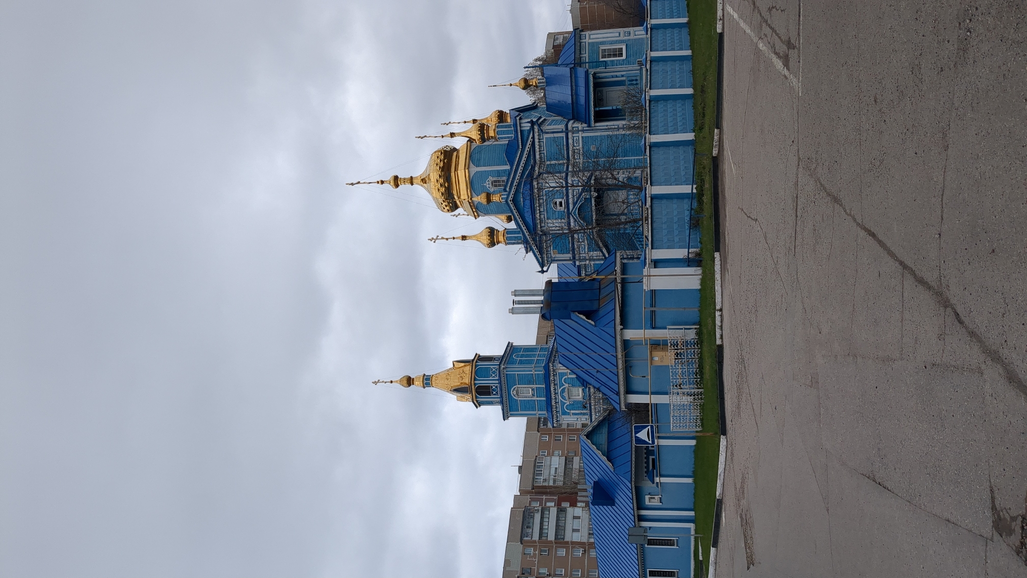 The image shows a vibrant blue wooden church with golden domes in Ulyanovsk, Russia. The church is adorned with intricate carvings and architectural details. Adjacent to the church is a grey asphalt parking area. A multi-story apartment building is visible in the background. The sky is overcast with grey clouds.