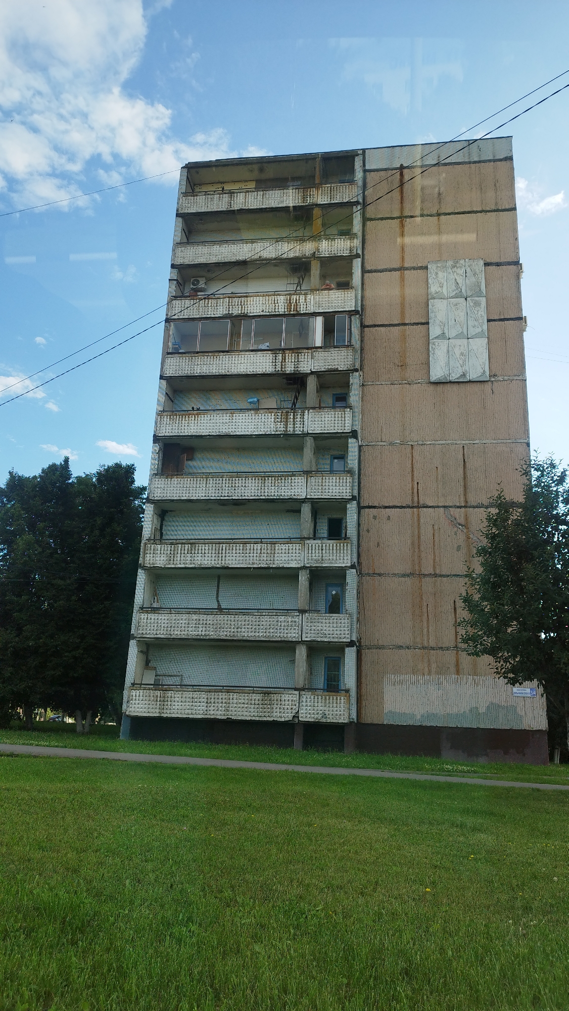 A tall, multi-story apartment building is depicted, with a visible residential apartment on the right side of the image. The building has multiple balconies with white patterned railings. The exterior walls are a pale beige concrete, with visible vertical stains and a large square panel on the upper right section. The building is surrounded by green grass and trees. The sky is blue with scattered white clouds. There are power lines running across the top of the image. A street sign visible on the building reads "СТАРООСКОЛЬСКАЯ 30", indicating the location is Starooskolskaya Street, building 30, in Stary Oskol, Russia.