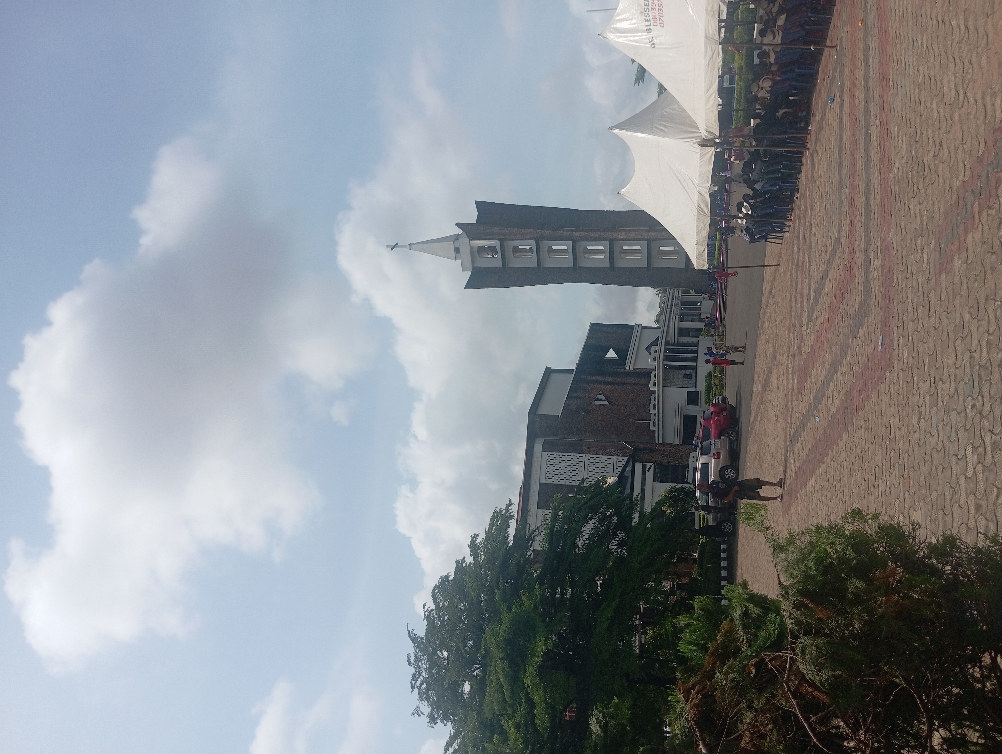 The image shows a building with a prominent bell tower under a cloudy sky. A large white tent is erected near the building, with rows of people sitting underneath it. The ground is paved with red and gray interlocking stones. There are trees and bushes in the foreground. Several vehicles are parked next to the building. The bell tower suggests this is the Basilica of the Most Holy Trinity in Onitsha, Nigeria. People are walking in the paved area.