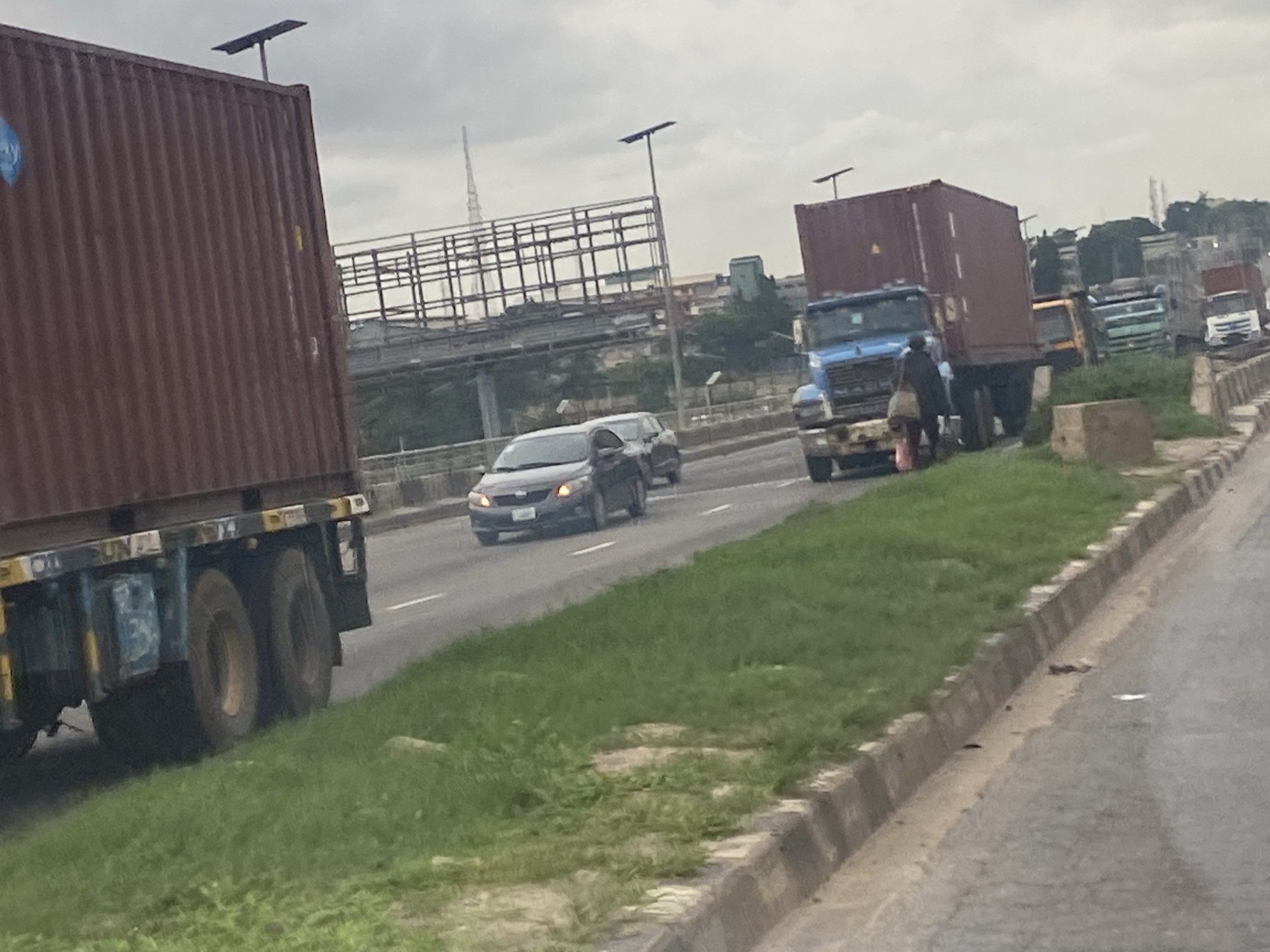 The image depicts a traffic scene on a highway, likely in Ikeja, Nigeria, with a cloudy sky overhead. In the foreground, a large red shipping container truck is partially visible on the left, with its wheels and lower chassis showing. To the right, a blue truck with a red shipping container is stopped, and a person in dark clothing carrying a light-colored bag is walking in front of it. Several other trucks, including a yellow truck, green and white trucks, and a white truck, are also part of the traffic jam, lined up on the right side of the road. A dark gray sedan is driving in the lane to the left of the blue truck. The road is bordered by a grassy embankment and a concrete curb. In the background, overhead bridges and structures can be seen, suggesting an urban or semi-urban environment. The overall impression is one of congestion and slow-moving traffic on a cloudy day.