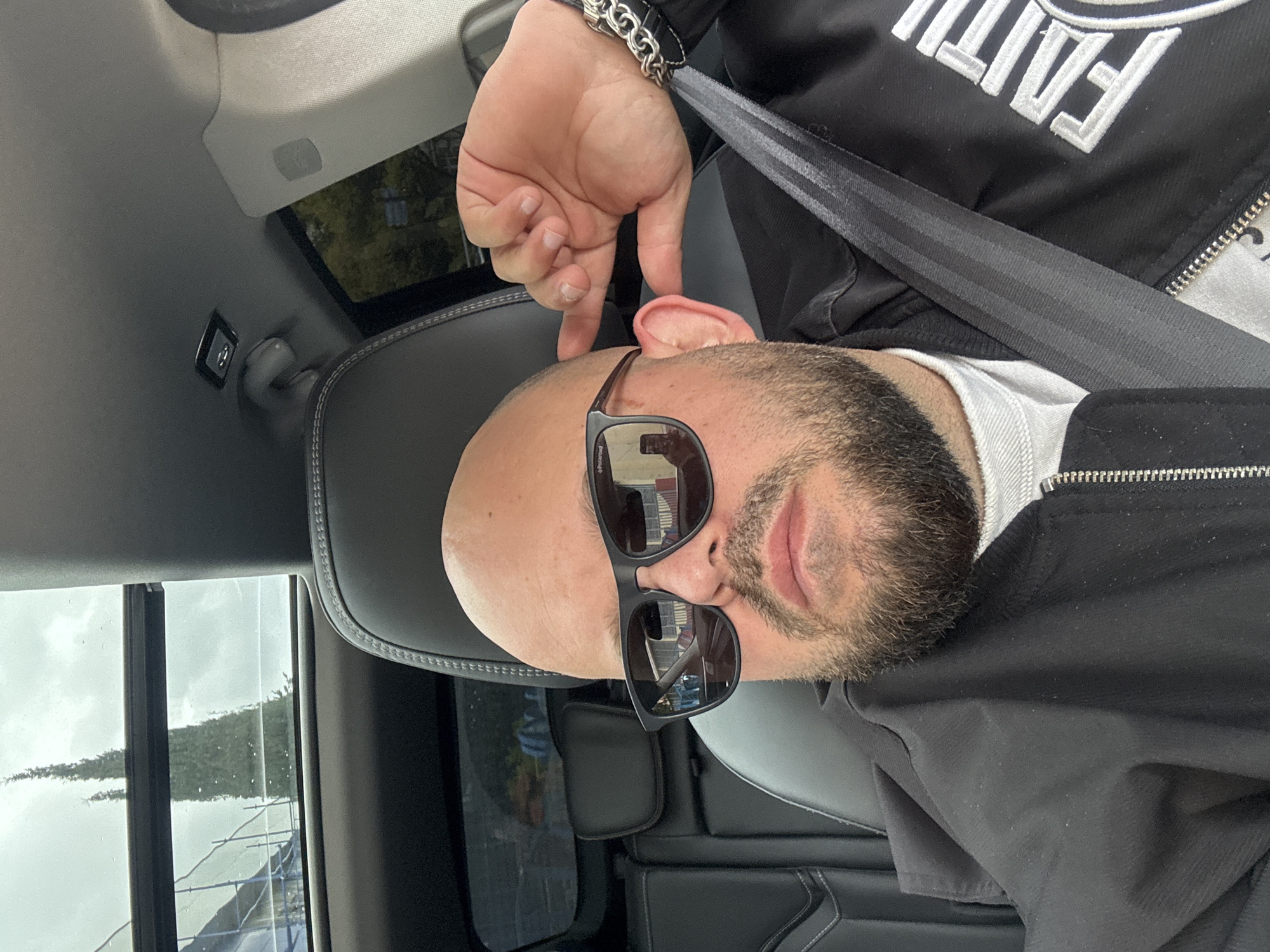 A man is in a car, wearing sunglasses and a black jacket with white embroidery, and a silver bracelet. He has a beard and a bald head. He's wearing a seatbelt. His fingers are near his ear. The location is not discernible as Sochi, Russia. The image does not have enough visible details to confirm the location.