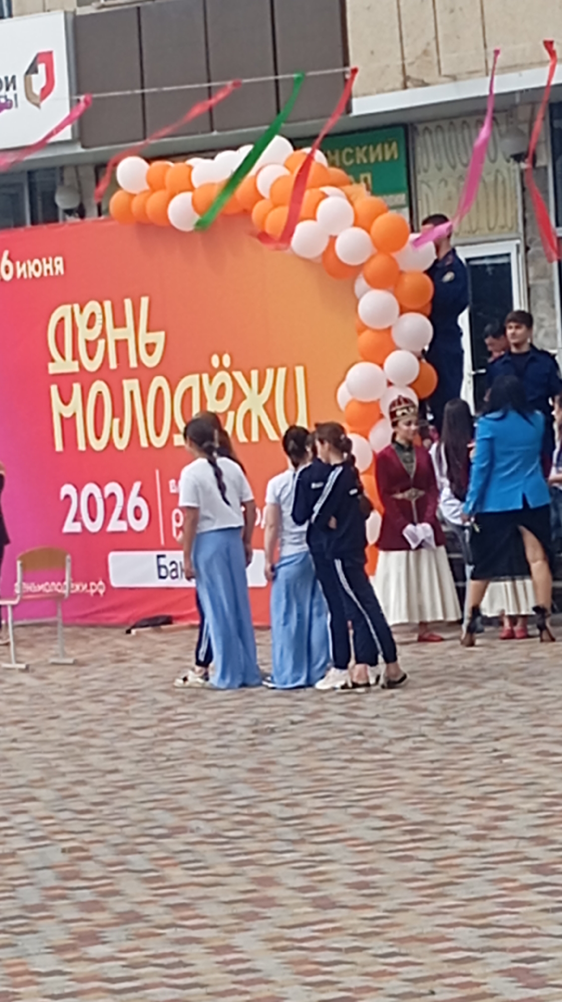 This media file captures an outdoor scene in Baksan, Russia, likely during a daytime event. The focal point is a large, vibrant banner that reads "ДЕНЬ МОЛОДЕЖИ 2026" in bold, yellow lettering against an orange and pink gradient background. This translates to "YOUTH DAY 2026," indicating a celebration of youth.
Several people are visible in the foreground, mostly young women and some men, dressed in a mix of casual and traditional attire. Some women are wearing long, flowing blue skirts and white tops, while others are in dark tracksuits. One woman is dressed in a striking red and gold traditional outfit, including a crown-like headpiece, suggesting a cultural performance or representation. Men in what appear to be uniforms, possibly security or official personnel, are also present, standing behind the banner.
The scene is decorated with a festive arch of orange and white balloons and colorful ribbons strung across the top. A paved ground, composed of interlocking bricks in shades of brown and beige, covers the area where the people are standing. In the background, a building with windows and signage can be seen.
The overall atmosphere appears to be one of celebration and gathering, likely for the "Youth Day" event. The lighting suggests it is daytime, and no specific weather conditions are discernible. Visible text includes the main banner, a smaller sign with the website "МОЛОДЕЖЬ.РФ", and partial text on the building in the background, one segment of which appears to be "НСКИЙ". The presence of traditional clothing alongside modern attire suggests a blend of cultural heritage and contemporary celebration.