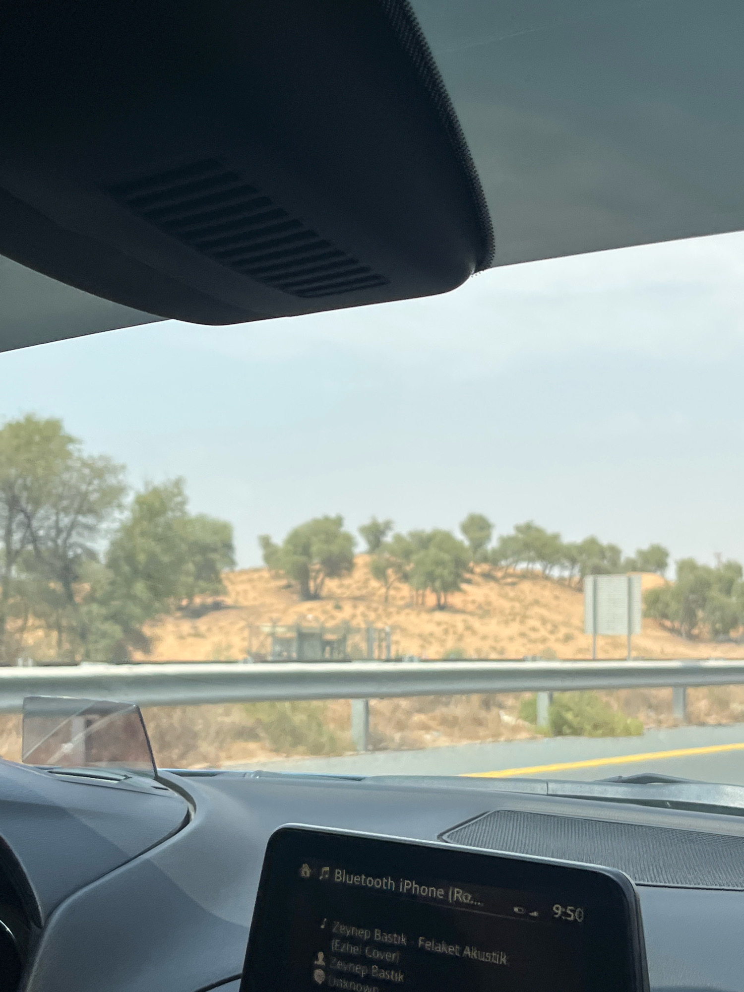 The image shows a view from inside a car in the city of Mohadhub, United Arab Emirates. The car's dashboard is visible in the foreground, with a screen displaying music information that reads "Bluetooth iPhone (Ro... Zeynep Bastik - Felaket Akustik". The time displayed on the screen is 9:50. Through the windshield, a semi-arid landscape with sparse vegetation and sandy terrain is visible. A highway barrier runs along the side of the road. The sky is bright and hazy, indicating daytime. The image is taken from the perspective of a passenger or driver.