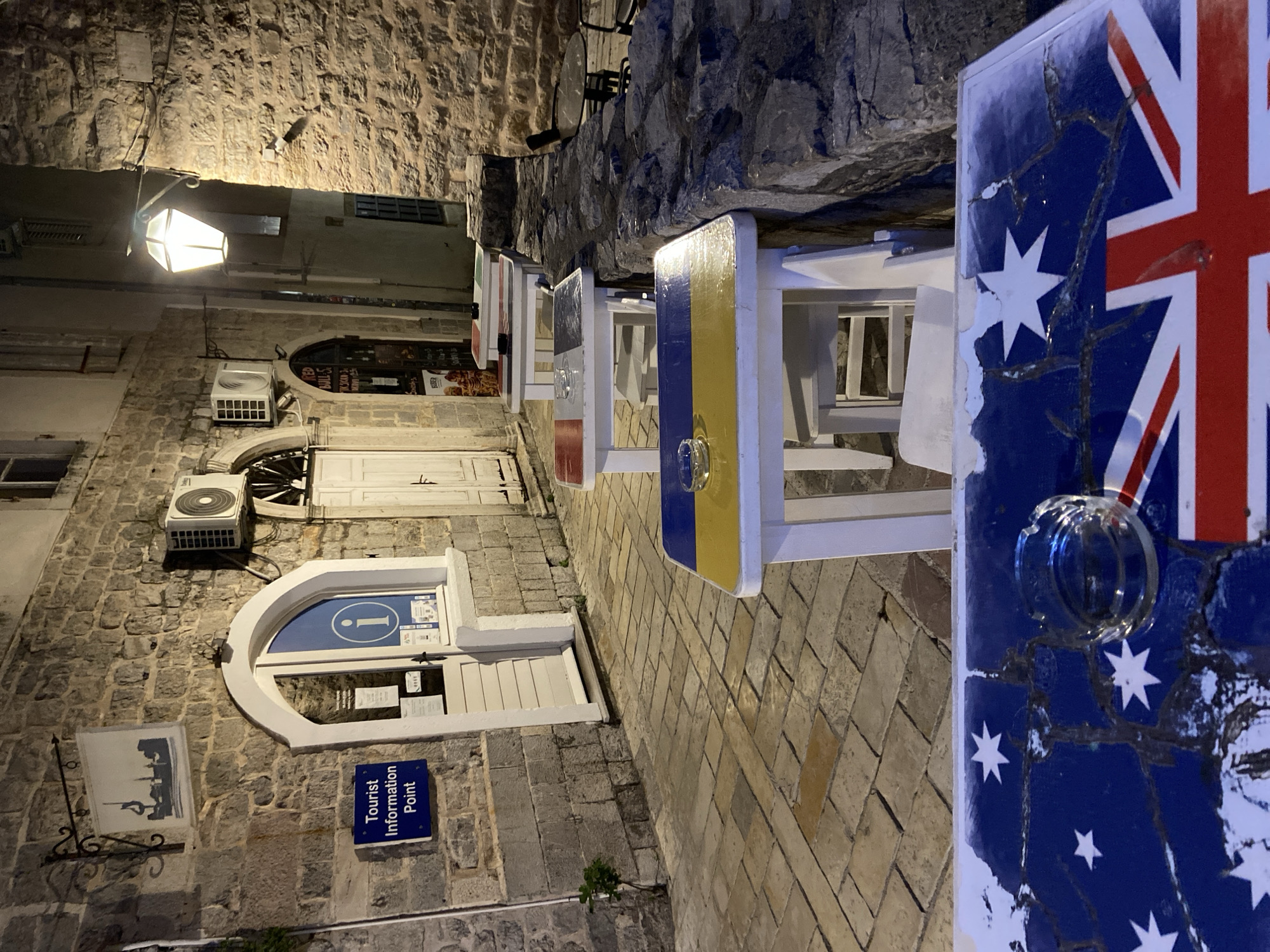 The image depicts a quiet, narrow street in an old town at night, likely in Budva, Montenegro. The cobblestone-paved street is lined with historic stone buildings illuminated by a traditional street lamp and other ambient light.
In the foreground and extending into the midground, a row of outdoor tables is arranged along the left side of the street. Each table's visible side is decorated with a national flag. The closest table clearly displays a weathered Australian flag, showing the Union Jack, the large white Commonwealth Star, and the Southern Cross constellation, with evident peeling paint and cracks. A clear glass or plastic holder is attached to its side. Further down the line, other tables feature flags resembling those of Ukraine (blue and yellow) and France (blue, white, red), suggesting an international theme or clientele.
The buildings on the left are constructed from rustic stone. Several details are visible on their facades, including two modern air conditioning units. A prominent arched entryway leads to a "Tourist Information Point," marked by a blue sign with white text stating "Tourist Information Point." Above this sign, an arched window or door features a circular blue sign with a white 'i' (information symbol). To the left of the information point, a wrought-iron bracket supports a rectangular sign displaying a dark silhouette, possibly of a local landmark or city skyline. Faint text like "RED BULL 5e" is visible through a nearby window, indicating a potential cafe or bar.
The scene is devoid of people, creating a peaceful and atmospheric evening ambiance. The artificial lighting and dark surroundings confirm it is nighttime.