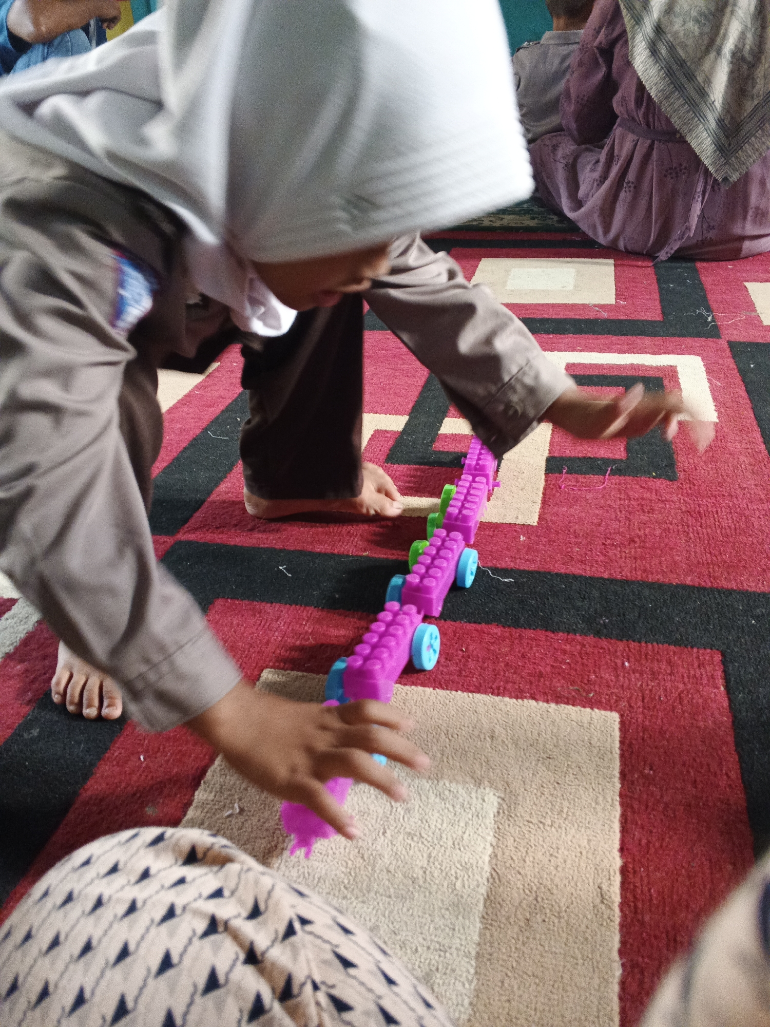 A child, wearing a white hijab and a brown uniform, is playing with a colorful toy train on a patterned rug in what appears to be a classroom or community space in Warungkiara, Indonesia. The child is hunched over, reaching out with their right hand towards the train, which consists of several magenta and green sections with blue wheels. Their left hand is also near the train. The child's feet are bare. In the background, another person is seated, wearing a purple dress and headscarf. The rug has a geometric pattern of red, black, and beige squares and rectangles. The lighting suggests an indoor setting.