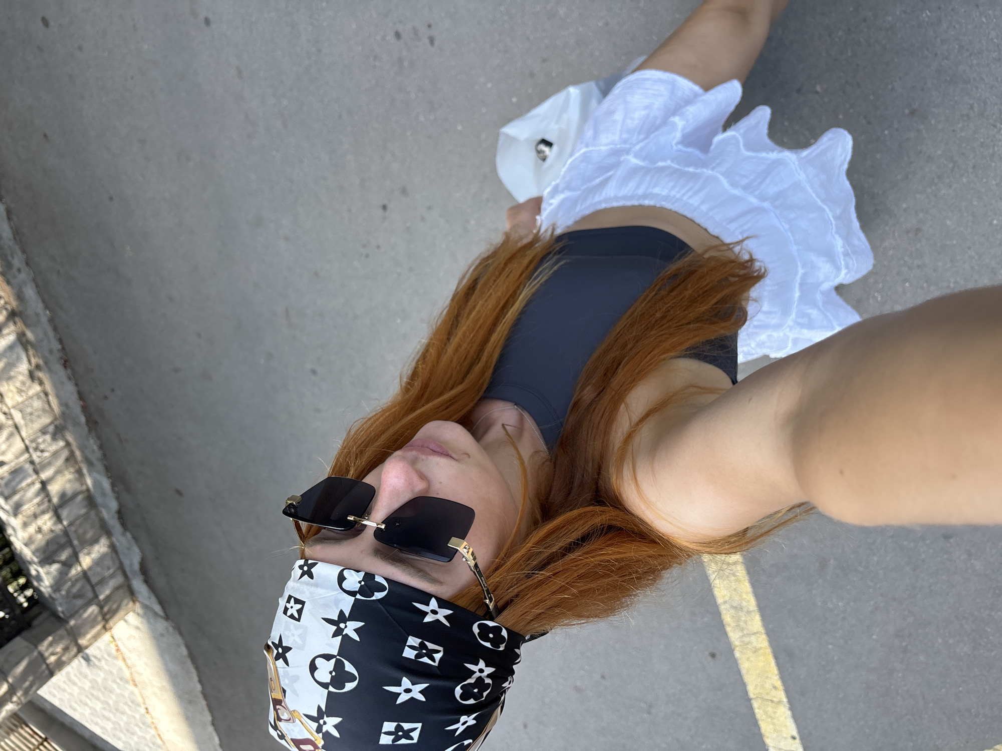 A high-angle, full-body selfie shows a woman with long, auburn hair wearing a black crop top, a white tiered ruffle skirt, and a black and white patterned bandana. She is also wearing large, dark-rimmed sunglasses. The woman is holding a white shopping bag in her right hand and is standing on a paved surface with a yellow line marking. In the background, there is a stone wall and foliage. The scene appears to be outdoors, possibly in an urban or park-like setting. The lighting suggests it is daytime.