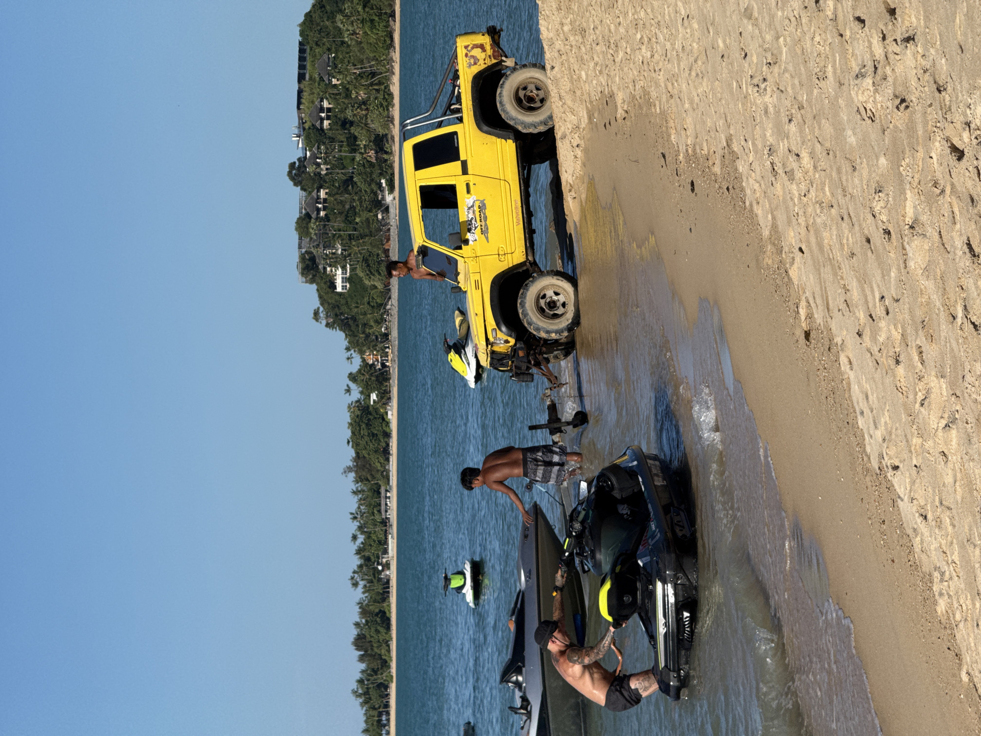In Baan Mae Nam, Thailand, a bright yellow off-road vehicle is parked on a sandy beach next to a body of water. The vehicle has a roll cage and a spare tire mounted on the back. The driver's side door is open, and a man is visible inside the vehicle. Several jet skis are present on the water and near the shore. Three men are interacting with the jet skis. One man is standing in the water near a jet ski, and another man is on the shore, looking at a jet ski. A third man, who is tattooed, is sitting on a jet ski. The scene appears to be set during the daytime, with clear blue skies. The beach is lined with lush green trees and buildings in the background, suggesting a tropical location. The overall mood seems to be relaxed and recreational, with people enjoying activities on the water. A sticker on the vehicle reads "OFF ROAD." Another visible text is "Valvoline" on the side of the yellow vehicle.
