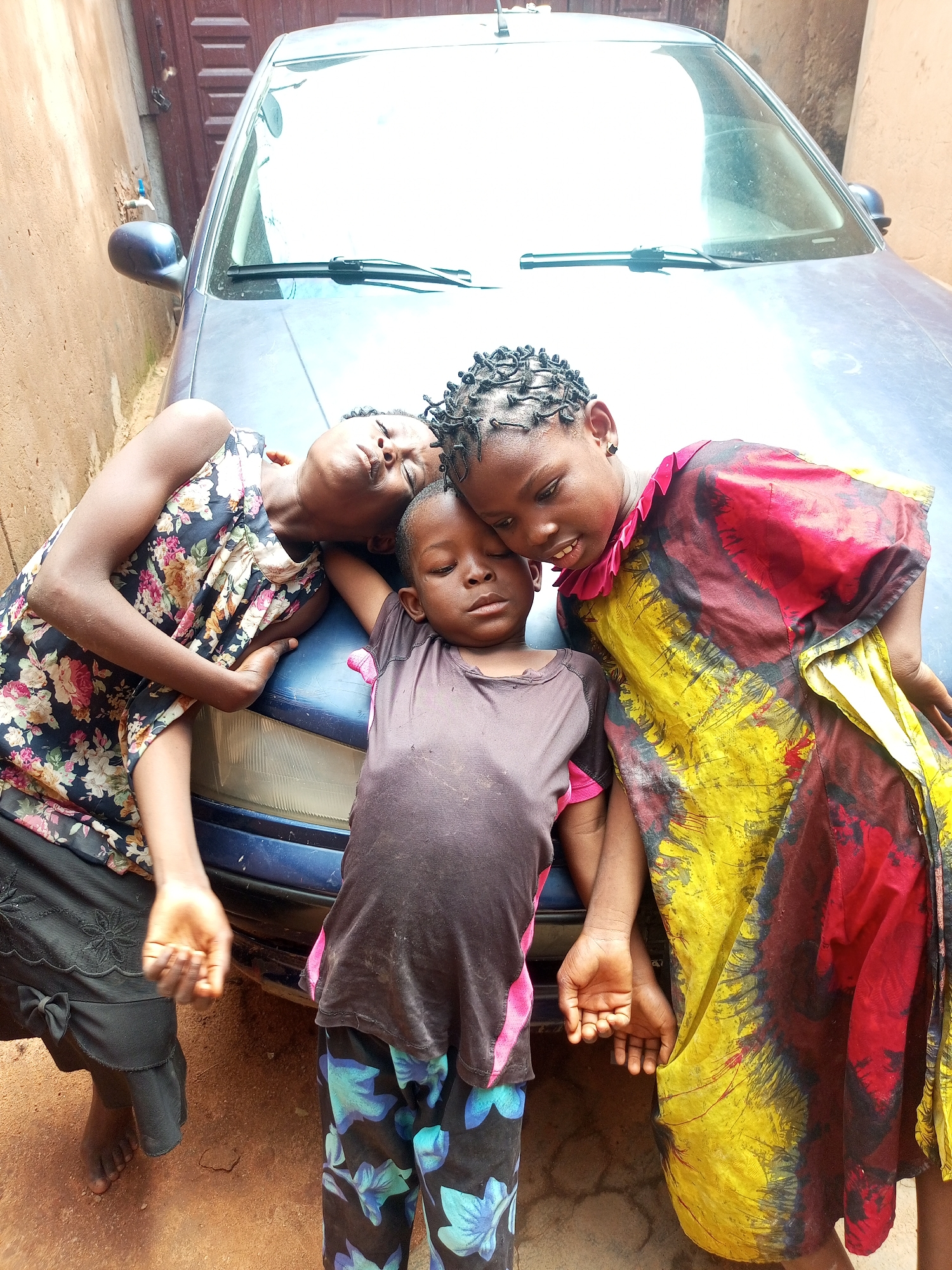 Three children, two girls and one boy, are lying on the hood of a blue car parked in an outdoor area that appears to be a residential courtyard in Tempure, Nigeria. The child on the left, a girl with dark hair, is wearing a black dress with a floral print. She is lying on her side, with her head resting on her arm and her eyes closed. The boy in the center is wearing a dark grey t-shirt and patterned pants. He is lying on his back, with his head tilted and his eyes closed. The girl on the right, with braided hair, is wearing a colorful dress with yellow, red, and black patterns. She is leaning over the other two children, looking down at them with her eyes open. Her right arm is resting on the boy's arm. The ground is covered in reddish-brown dirt. A brick wall is visible in the background, along with a wooden door. The car's windshield and hood are prominent in the foreground.