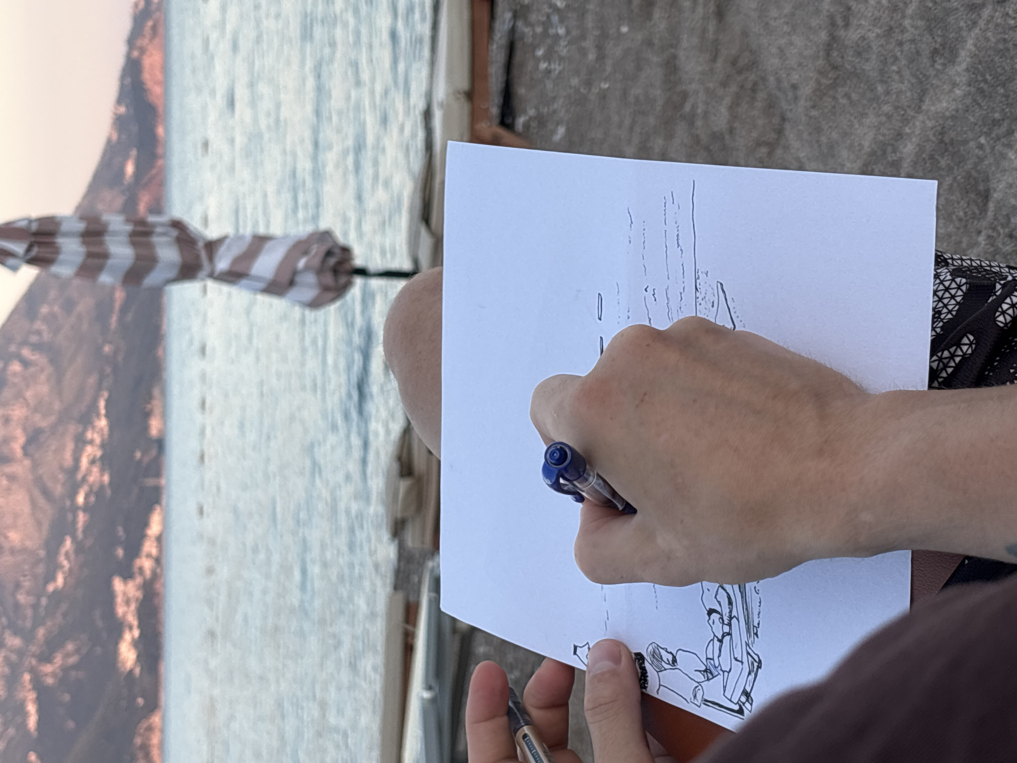 A person is drawing on a sheet of white paper with a blue pen, sitting on a beach. The drawing depicts two figures on a beach chair. The background shows the sea, a closed umbrella, and hills in the distance. The scene is set in Bečići, Montenegro. The lighting suggests it might be late afternoon or early evening. The person is wearing patterned shorts.