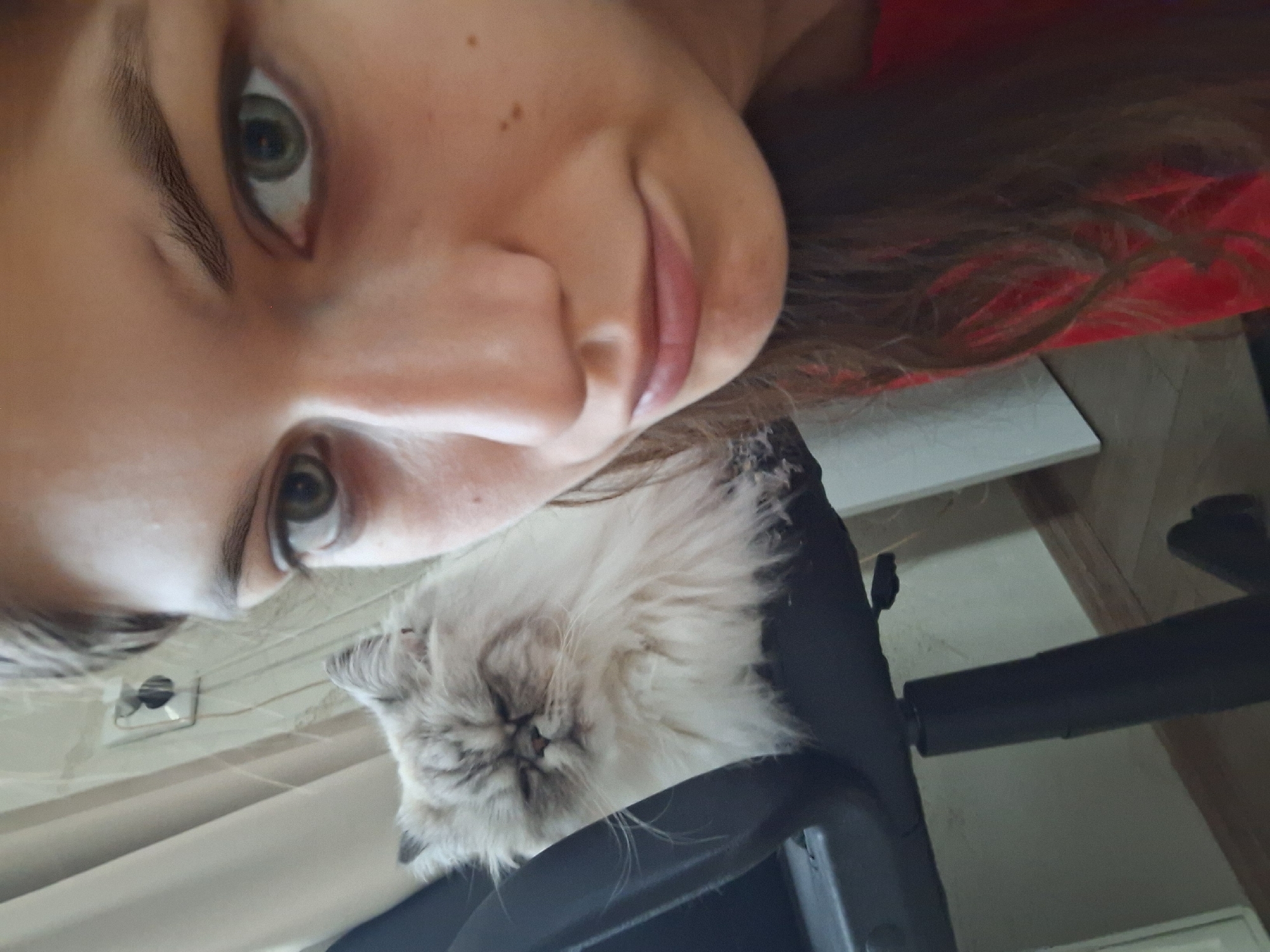 A woman is looking down into the camera with a slight smile. Below her is a cat, possibly a Persian or Himalayan breed, sitting in what appears to be a black office chair. The background includes a wall, possibly paneled or striped. The location is not possible to determine based on the available information.