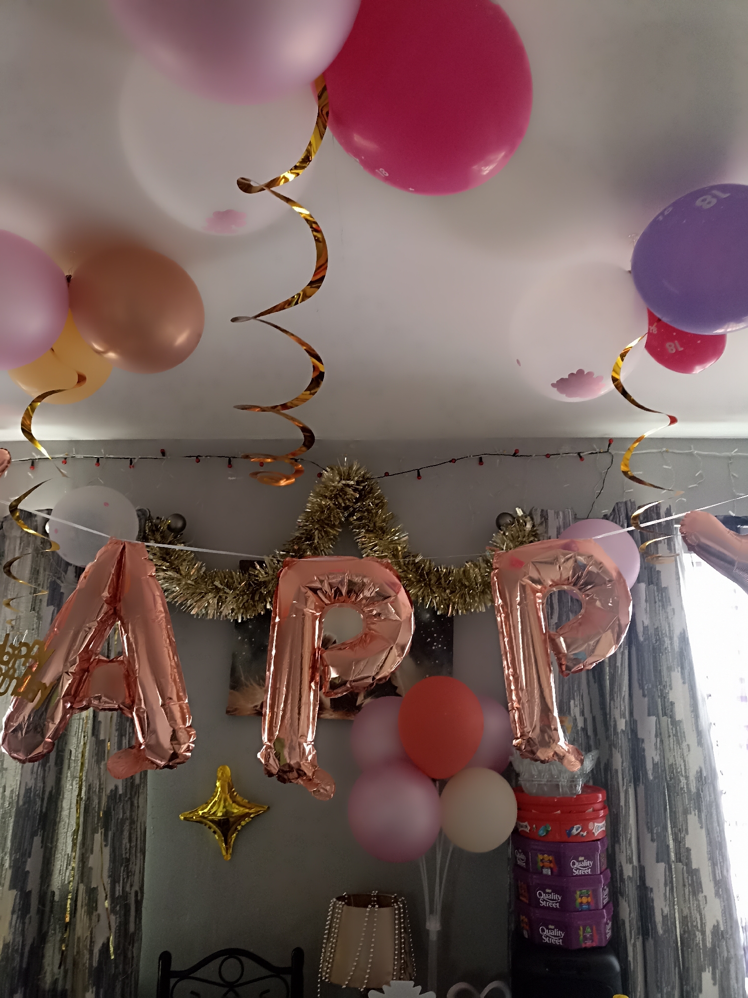 The image depicts a room decorated for a celebration, likely a birthday. The ceiling is adorned with various balloons in shades of pink, peach, purple, and white. Some balloons are printed with the number "18". Gold and silver streamers hang from the ceiling, adding a festive touch.
Dominating the lower portion of the image are large, rose gold foil balloons spelling out "APP". These balloons are arranged to be the centerpiece of the decoration. Below the "APP" balloons, there is a stack of boxes that appear to be chocolates, specifically "Quality Street" brand. To the left of the "APP" balloons, a smaller gold star-shaped balloon is visible.
In the background, there are grey curtains with a patterned design. A string of small red lights is also visible along the wall. A portion of a dark metal chair is visible at the bottom of the image.
The scene suggests a party environment within a domestic setting. There are no individuals clearly visible in the image. The lighting appears to be a mix of ambient room light and possibly light from a window to the right. The overall impression is one of a cheerful and decorated space ready for a special occasion.