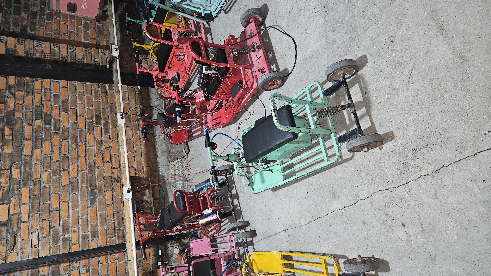 The image captures an indoor scene featuring multiple small, custom-built recreational vehicles, resembling go-karts or electric scooters, parked on a concrete floor. On the left, a textured brick wall with a dark horizontal beam spans the length of the image. White electrical conduits with visible outlets are mounted vertically on the brick wall, suggesting a utility or storage area.
There are at least five such vehicles, distinct in color. The most prominent one in the upper middle is red, featuring a metal frame, a black seat, exposed wiring, and rubber wheels. Below it, a light blue or mint green vehicle, also with a black seat and exposed components, appears to be connected to the red one by wires. Further down, parts of a pink and a yellow vehicle are visible. All vehicles exhibit a robust, somewhat industrial design with visible suspension springs, steering mechanisms, and numerous wires, suggesting they are electric-powered or perhaps undergoing maintenance/charging. Some vehicles show signs of use, with scuff marks on wheels and frames.
The concrete floor displays cracks, indicating wear. No people are present, and no specific activity is clearly depicted beyond the vehicles being parked. The lighting is even, suggesting an indoor setting or diffused light, so the time of day and weather are not discernible. No clear, legible text is visible anywhere in the image. The scene suggests a workshop, garage, or storage facility for these vehicles, located in Cam Đức, Vietnam.