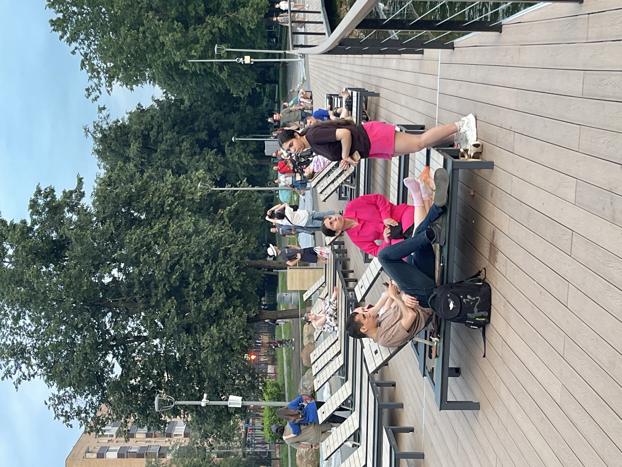 The image depicts a park scene with people relaxing on lounge chairs on a wooden deck. In the foreground, a man in a brown shirt and jeans is seated on a lounge chair, looking at his phone. A black backpack is placed on the deck next to him. To his right, a woman in a bright pink jacket is seated, and a younger woman in a dark top and pink shorts stands beside her. In the background, more people are visible on lounge chairs and walking around. The environment includes trees, a building in the distance, and a body of water with a railing along the deck. The overall atmosphere suggests a leisurely day in a public park. The presence of a traffic light in the background, along with the architectural style of the building, suggests an urban park setting.
