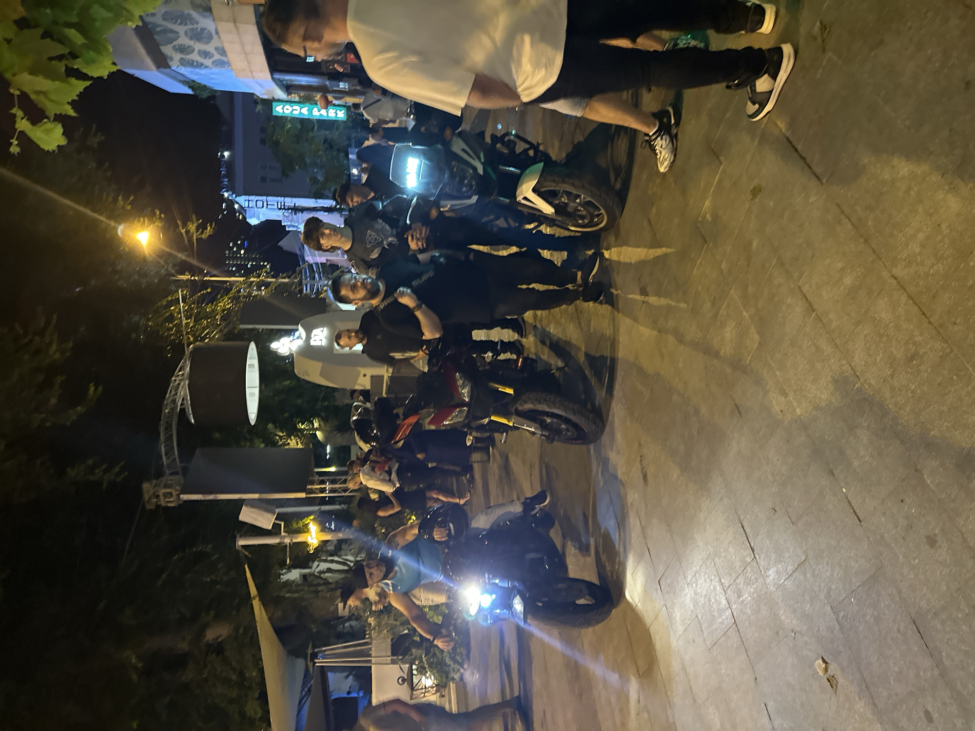 The image depicts an outdoor scene at night in the city of Odesa, Ukraine, likely on a pedestrian walkway or street lined with businesses and trees. A group of people are gathered, some of whom are standing near or on motorcycles.
In the foreground, a man in a turquoise tank top and a black beanie is seated on a black motorcycle, facing left. The motorcycle's headlight is illuminated, casting a bright light forward. He holds a black helmet in his left hand. To his right, another motorcycle, predominantly black with red accents and yellow suspension forks, is parked.
Further back and to the right, two men are standing next to a white and green motorcycle. One man, wearing a black t-shirt and pants, has a beard and is holding a black strap over his shoulder. The other man, younger, is wearing a dark patterned t-shirt and dark pants. Behind them, a man with a distinctive haircut and dark clothing is standing. To his right, another individual in dark clothing is visible.
In the mid-ground, a crowd of people are milling about. A man in a white t-shirt and shorts is standing on the far right, facing away from the camera, with a man in black pants and white sneakers standing in front of him. Behind the main group, several individuals are visible, including women in dresses, suggesting a social gathering or nighttime entertainment venue.
The environment is lit by streetlights and illuminated signs. One sign, vertically oriented and glowing green, reads "AQUA PARK". Trees with foliage are visible overhead, and architectural elements of buildings can be seen in the background, suggesting an urban setting. The ground is paved with large, square stone tiles, with shadows cast by the people and objects. The overall impression is of a lively evening scene in a public urban space.